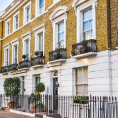 A row of elegant, yellow-bricked townhouses with white accents and black wrought iron railings. The buildings feature large windows, potted plants near the doors, and small balconies. A speed limit sign showing 20 km/h is visible on the sidewalk.