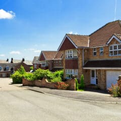 A suburban street with several two-story brick houses featuring tiled roofs and attached garages. The homes are surrounded by well-maintained gardens, and the sky above is clear and blue.