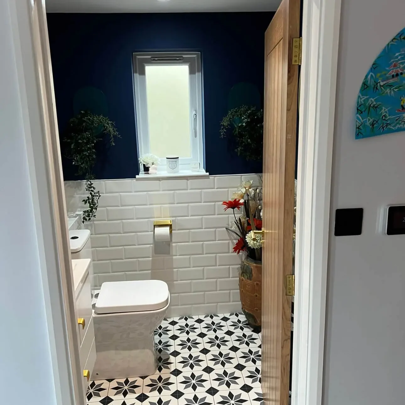 Small bathroom with navy blue walls and white subway tile wainscoting. The floor features black-and-white geometric patterned tiles. There is a white toilet, a small vanity on the left, a narrow window, and decorative plants and flowers.