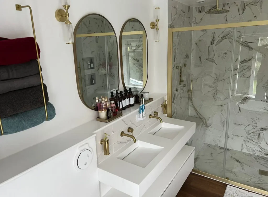 Modern bathroom with a double sink vanity featuring gold faucets and two oval mirrors. Several bottles and toiletries are neatly arranged between the sinks. To the right, a glass-enclosed shower with marble-patterned tiles and gold fixtures.