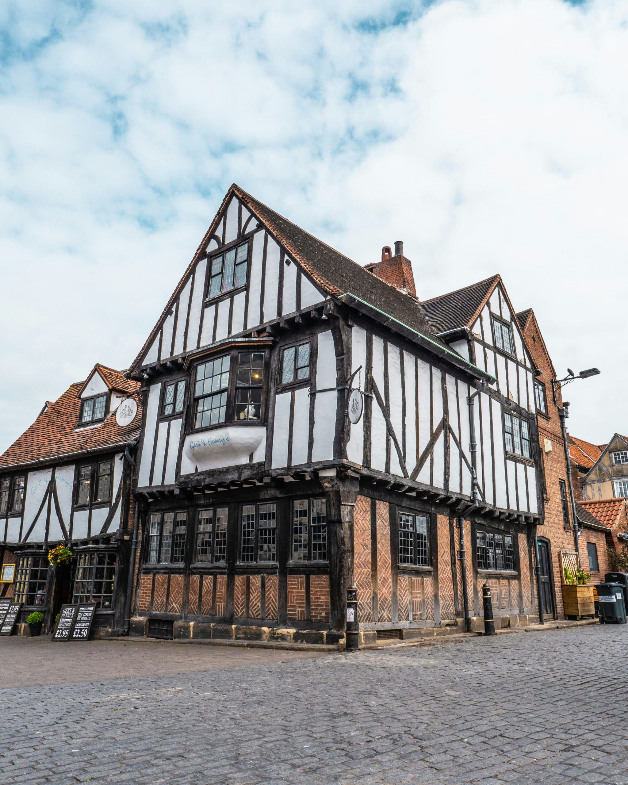 An old, timber-framed building with white walls and black wooden beams sits at a cobblestone street corner. The picturesque structure features multiple gabled roofs, leaded windows, and a sign hanging on the front facade. The sky is partly cloudy.