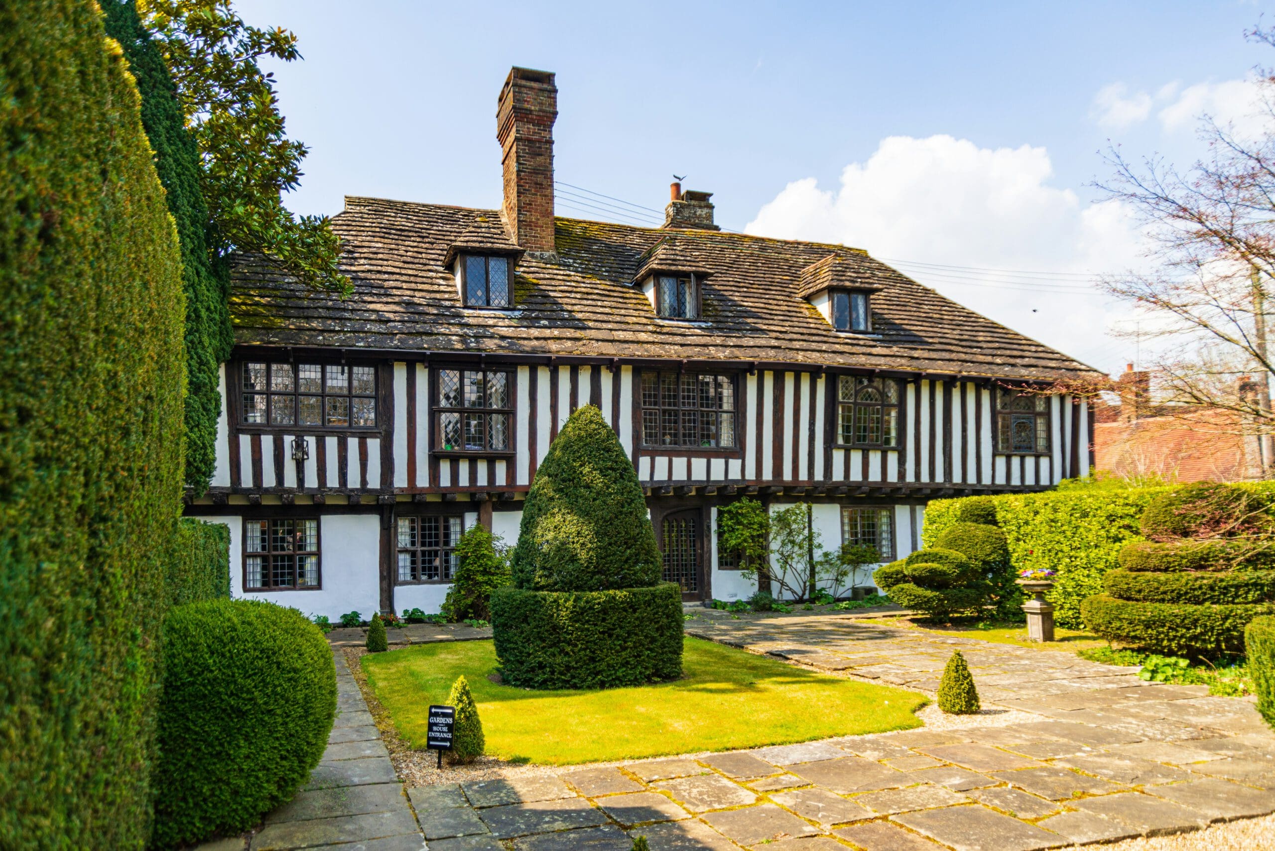 A historic Tudor-style house with a thatched roof, wood and plaster facade, and brick chimney. The front yard features manicured hedges, shaped topiaries, and a stone pathway leading to the entrance. The scene is set under a clear sky with a few clouds.