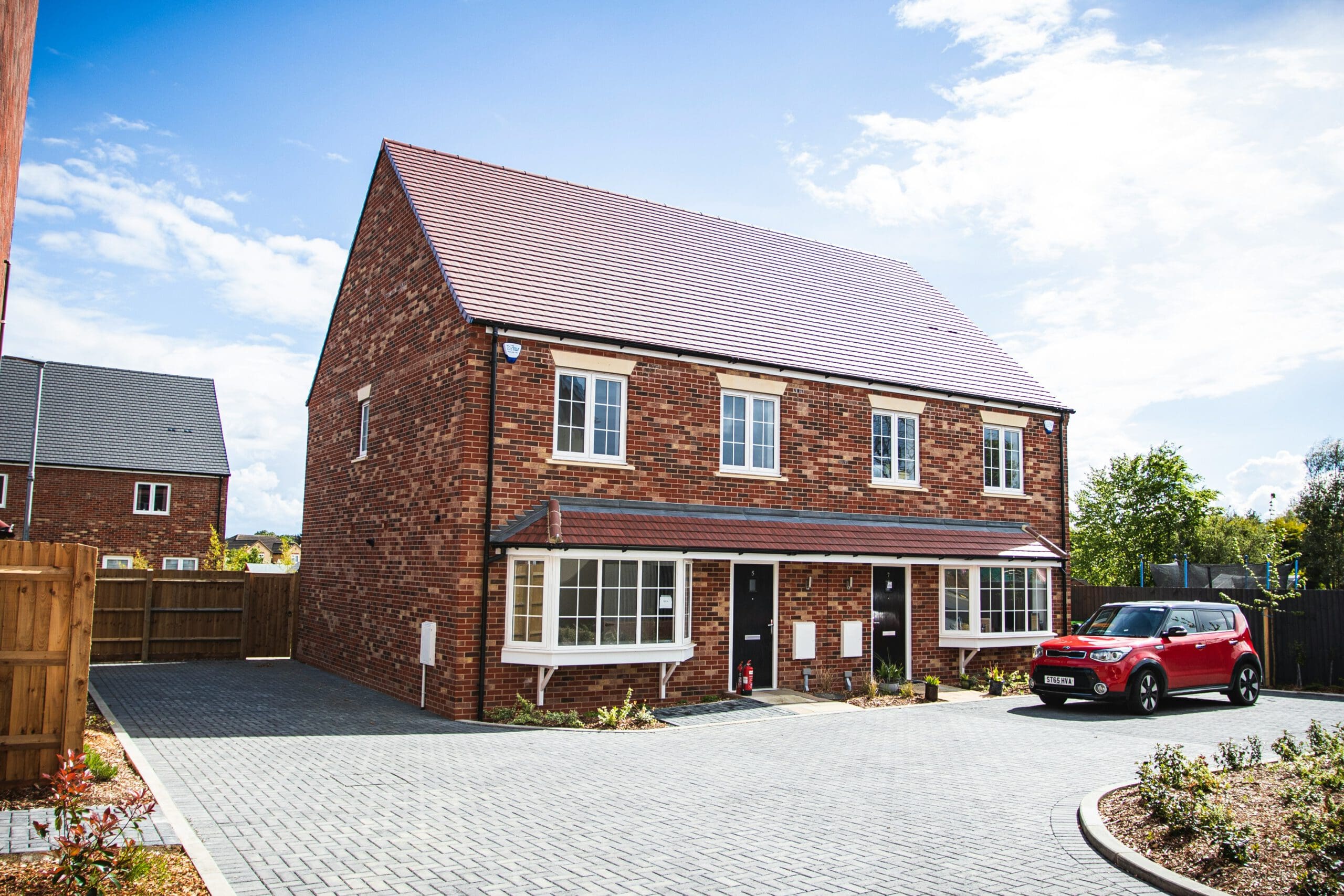 A red brick semi-detached house with a tiled roof, featuring two front doors and bay windows. A red car is parked on the paved driveway in front of the house. The sky is bright with scattered clouds, and there are other houses and trees in the background.