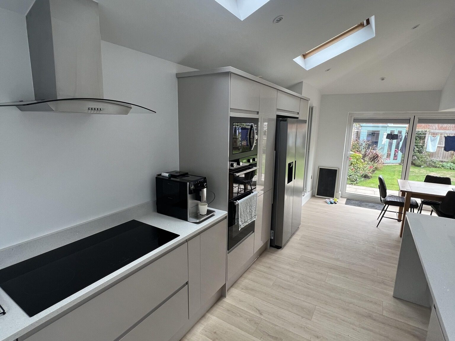 Modern kitchen with white cabinets and countertops, featuring a built-in oven, microwave, and refrigerator. An electric stovetop with a hood is on the left. A coffee machine is next to the oven. The far end opens to a dining area with a sliding door to the garden.