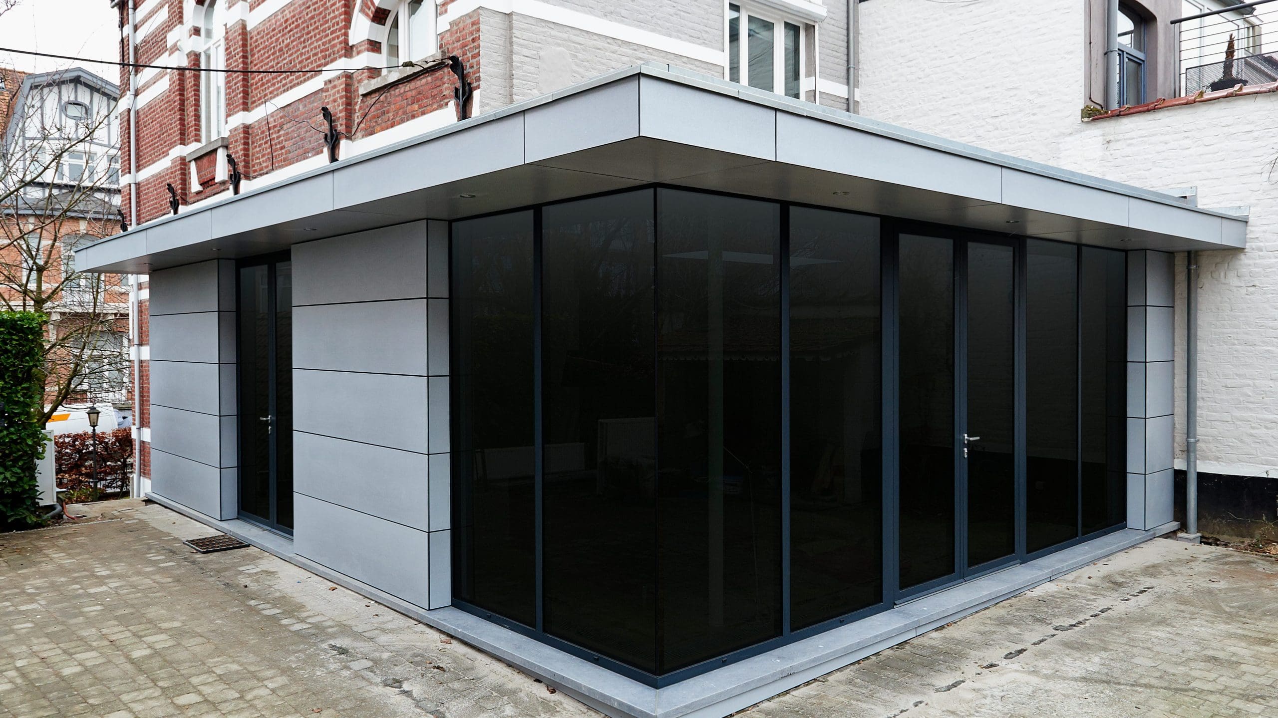 A modern extension with large dark-tinted glass windows and sleek grey cladding adjoins a traditional brick building. The area around the extension is paved with light-colored bricks. The scene is outdoors with some greenery visible on the side.