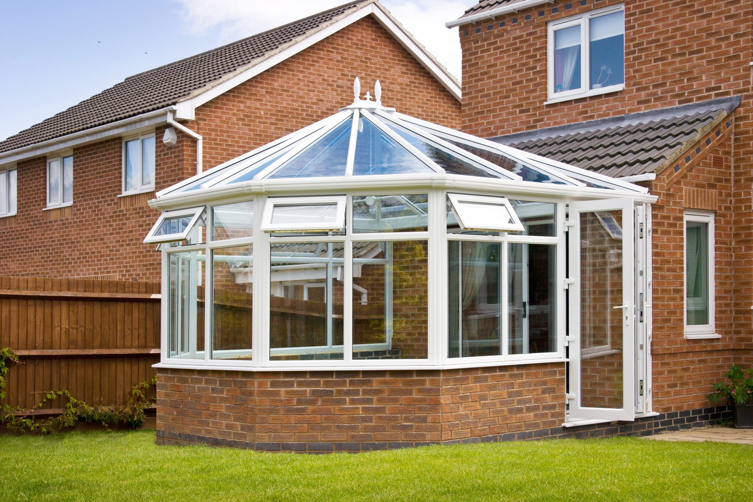 A modern brick house with a white-framed glass conservatory extending from the side. The conservatory has large glass windows and a pitched glass roof. The house and conservatory are surrounded by a well-maintained green lawn with a wooden fence in the background.