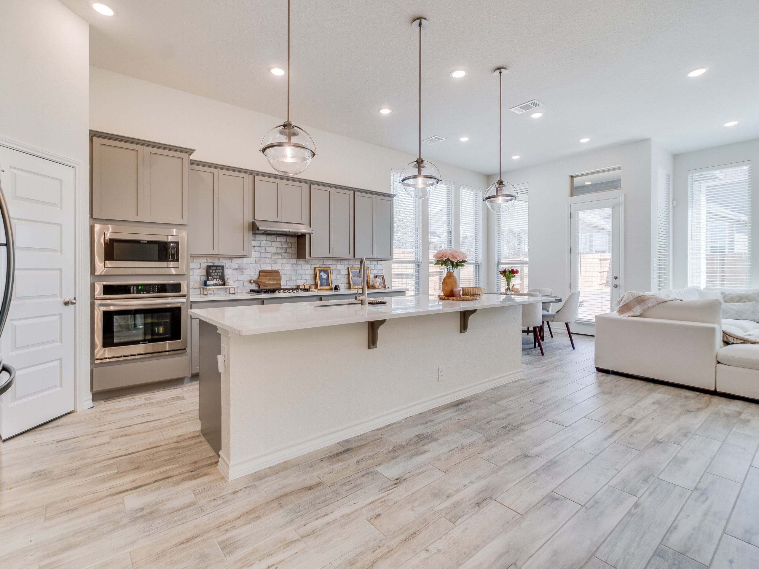 A modern, spacious kitchen and living area in a house with light wood flooring. The kitchen has beige cabinets, stainless steel appliances, a large island with white countertops, and pendant lights. The living area features a large white sectional sofa. Ample natural light fills the space from large windows.