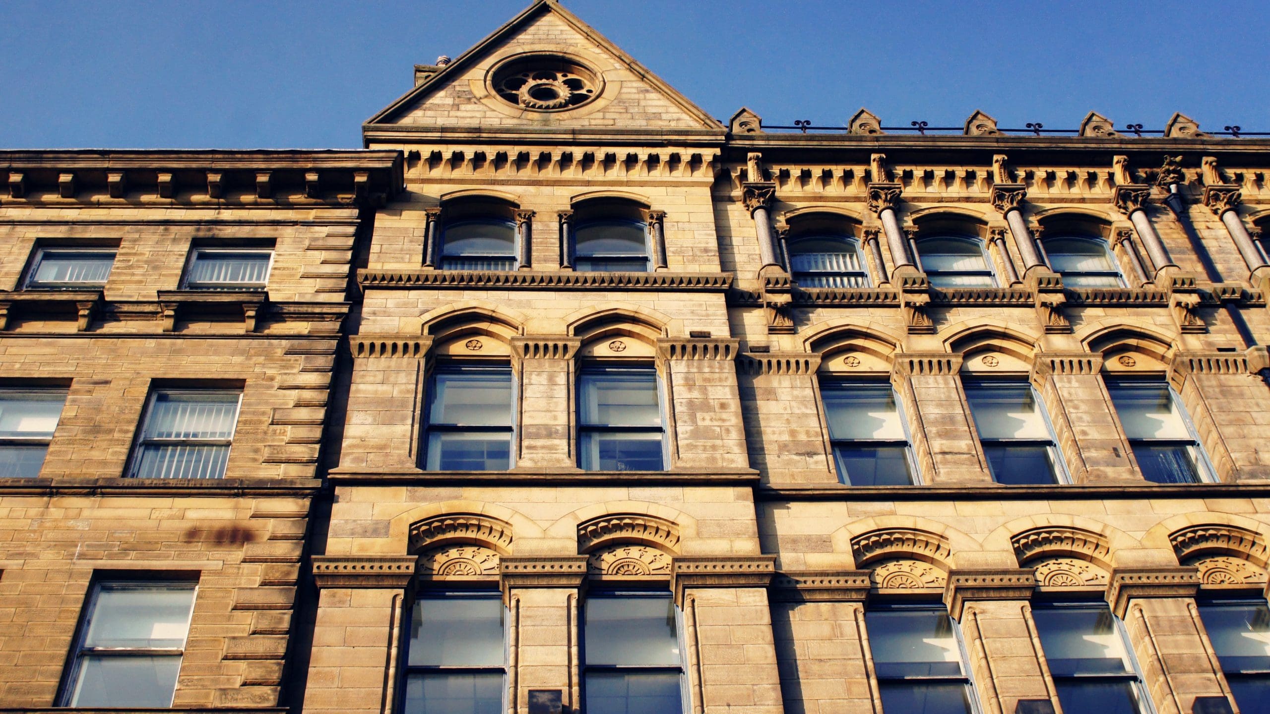 A close-up view of a historic multi-story stone building with tall windows and ornate architectural details under a clear blue sky. The facade features arched windows, intricate cornices, and a triangular gable with a circular decorative element.