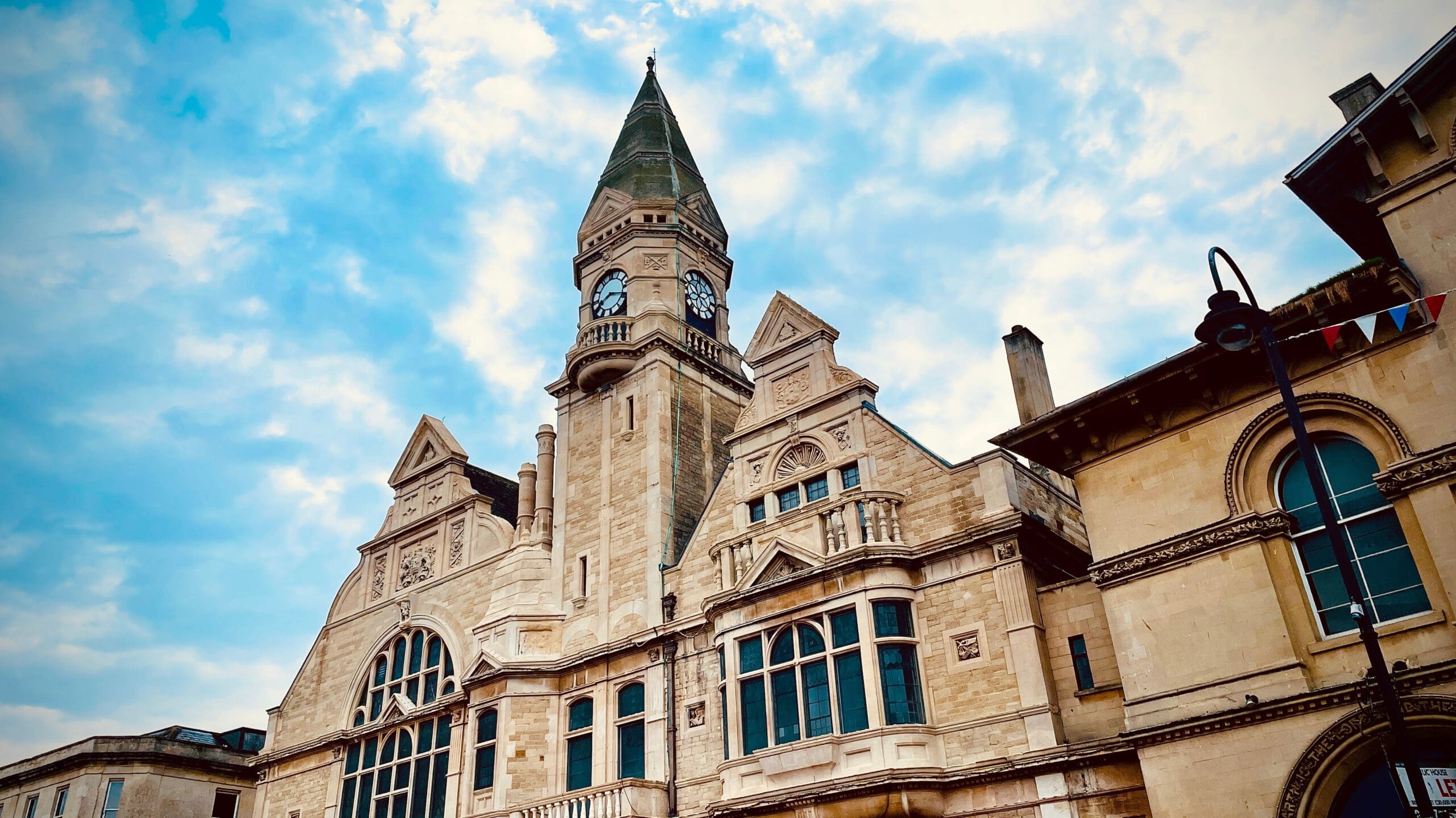 A historic building with a clock tower under a partly cloudy sky. The structure features large arched windows, ornate stonework, and various architectural details. A streetlamp with bunting is visible on the right side of the image.