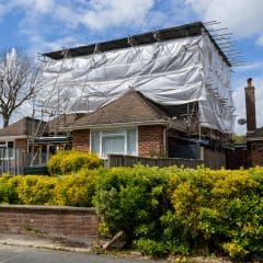 A single-story brick house under renovation is partially covered in scaffolding and white protective sheeting. The surrounding garden is well-maintained with green bushes. Other houses and trees are visible in the background under a partly cloudy sky.