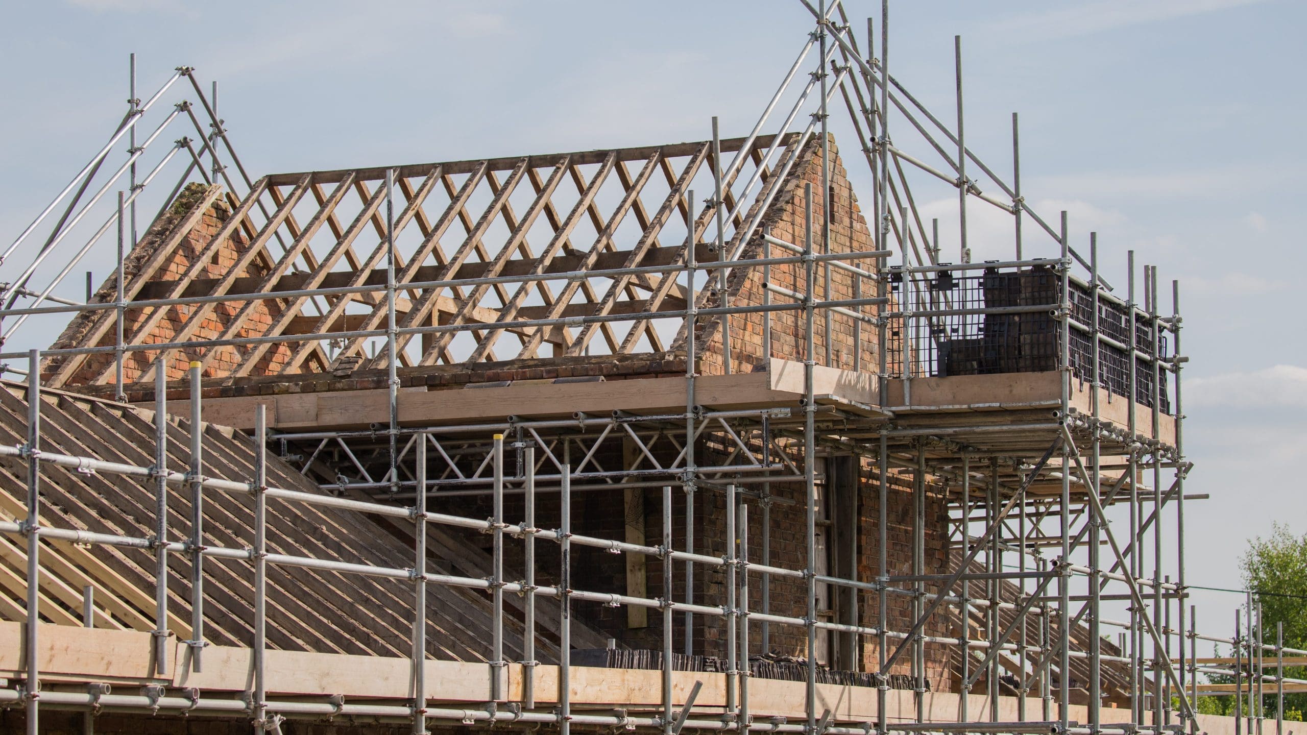 A partially constructed or renovated house with exposed brick walls and wooden roof trusses. The site is surrounded by metal scaffolding, providing support and access for workers. The sky is clear and blue.