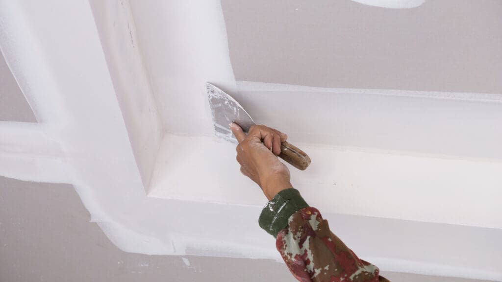 A person wearing a camouflage-patterned shirt uses a putty knife to apply white drywall compound to the seams of a ceiling. The image focuses on the task being performed, highlighting the hand and the tool.