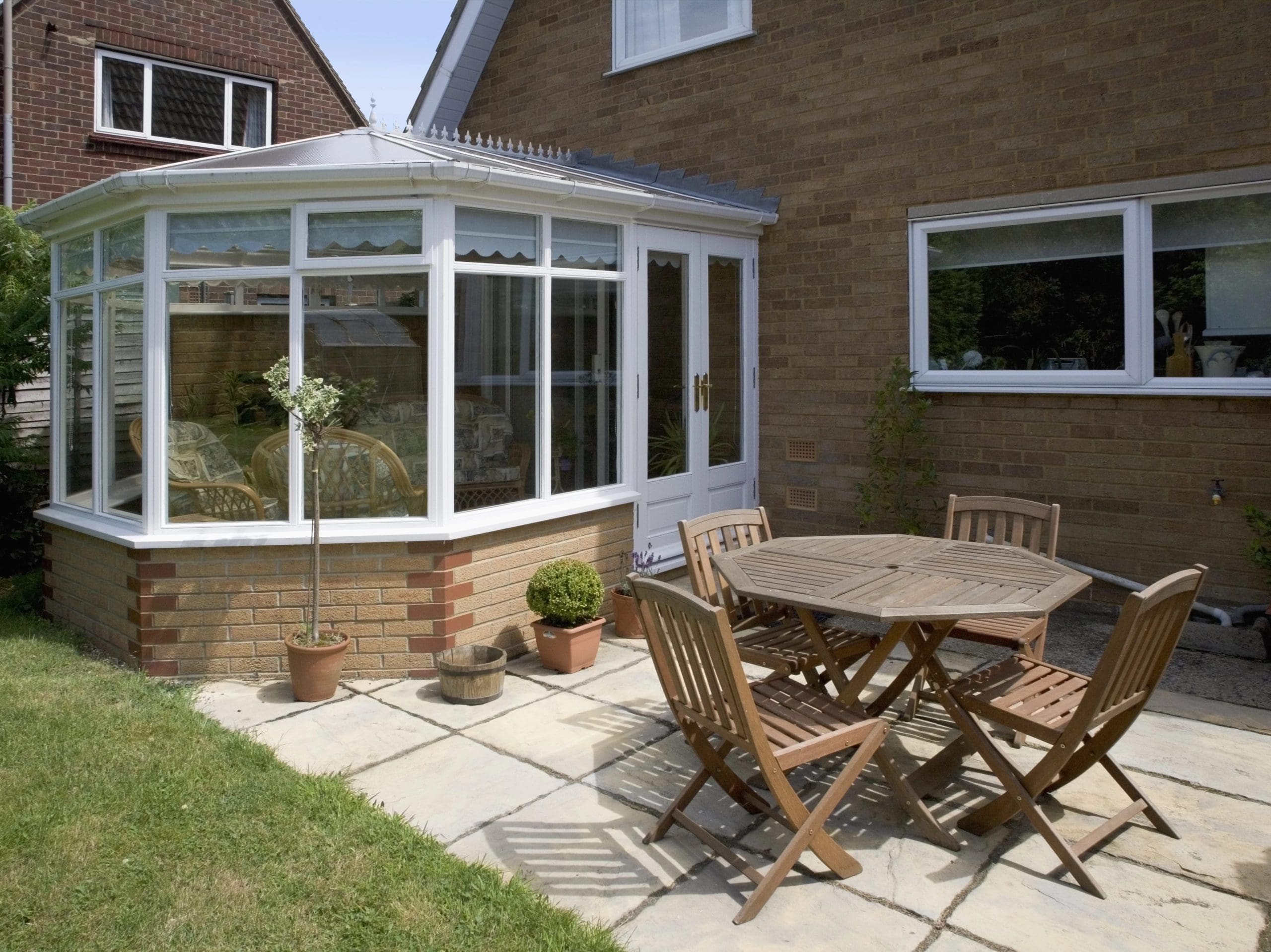 A brick house with a white-framed sunroom featuring large windows is seen. Adjacent to the sunroom, on a paved patio, is a wooden outdoor table with six matching chairs. Several potted plants are placed around the patio area, and a portion of the lawn is visible.