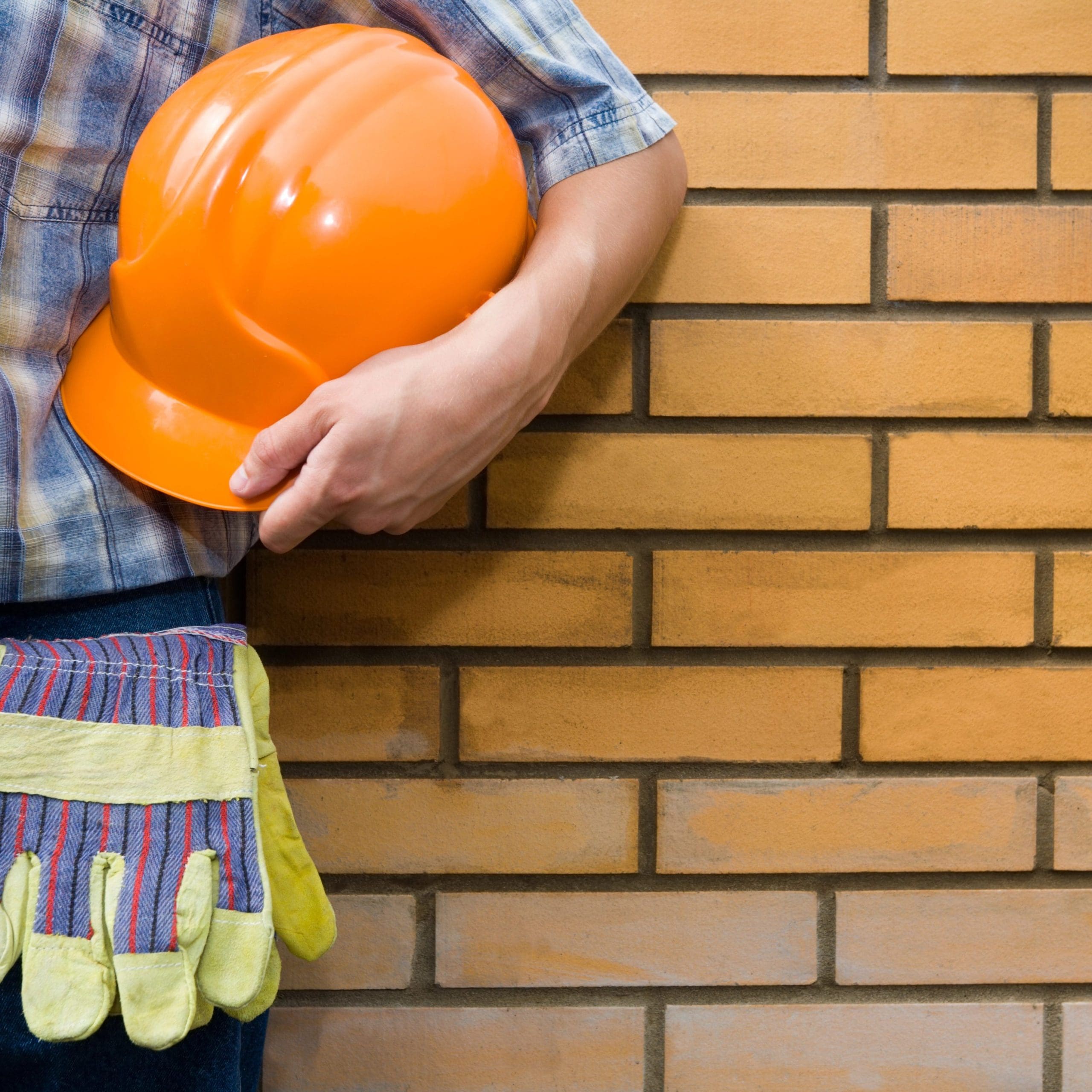 A person in a plaid shirt stands against a brick wall, holding an orange safety helmet and wearing a tool belt with gloves hanging from it.