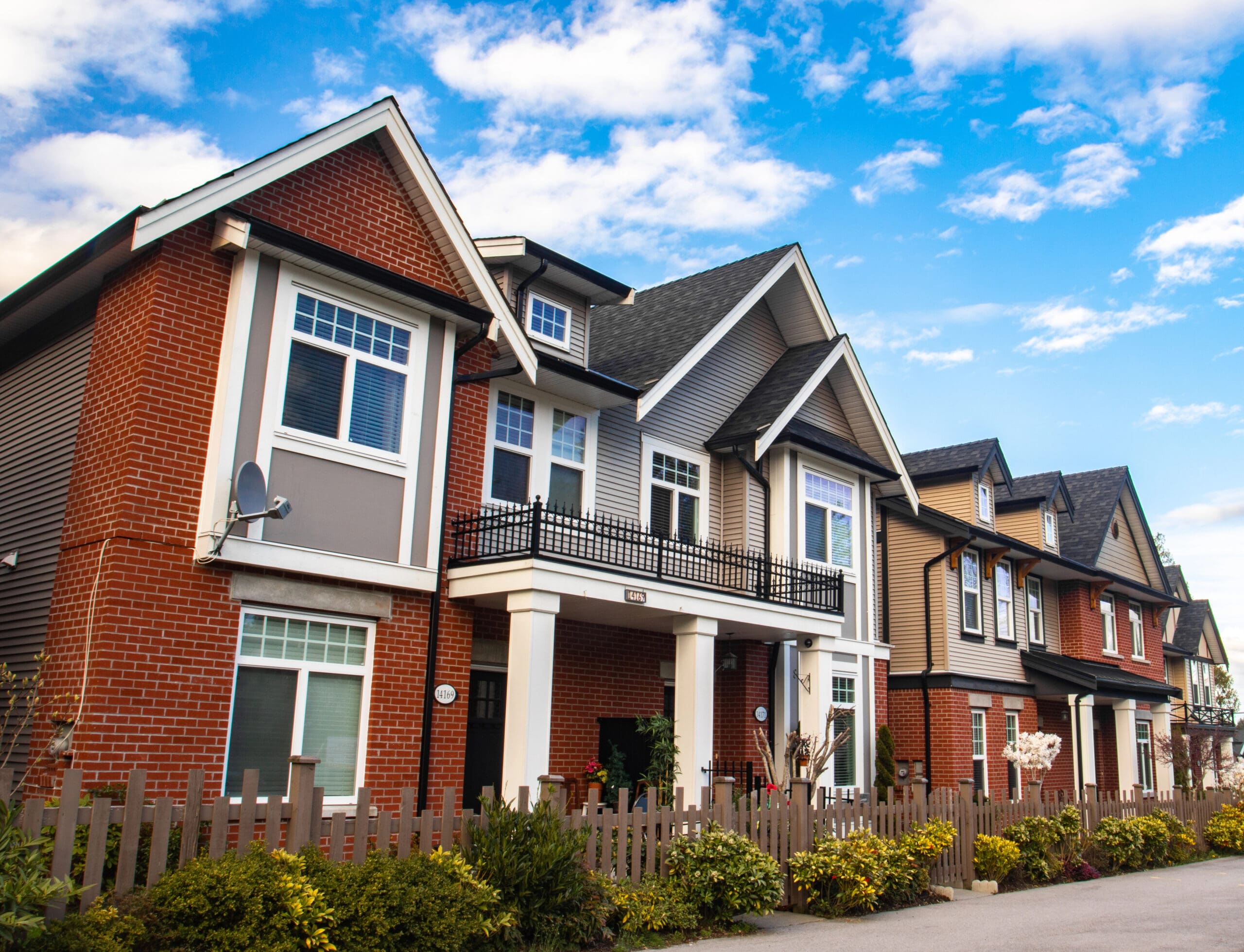 A row of modern suburban houses with varying facades, including brick and siding. Each home features large windows and gable roofs. A small, well-kept front garden and a wooden picket fence line the front of each property. The sky is bright and partly cloudy.