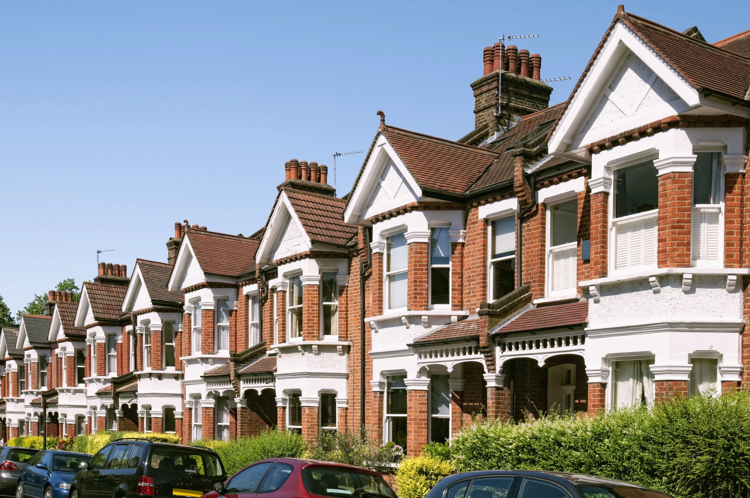 A row of traditional two-story red brick houses with white accents and bay windows. The homes feature gable roofs and chimneys. Parked cars are visible along the street in front, and a hedge lines the front yards. The sky is clear and blue.