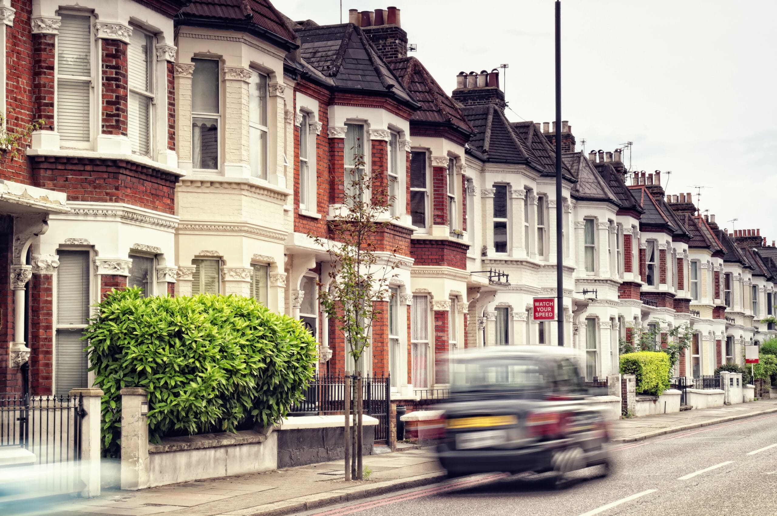 A row of red-brick Victorian terraced houses with white accents lining a street in a residential neighborhood. A black London taxi speeds by in the foreground, with a few small trees and parked cars along the sidewalk.