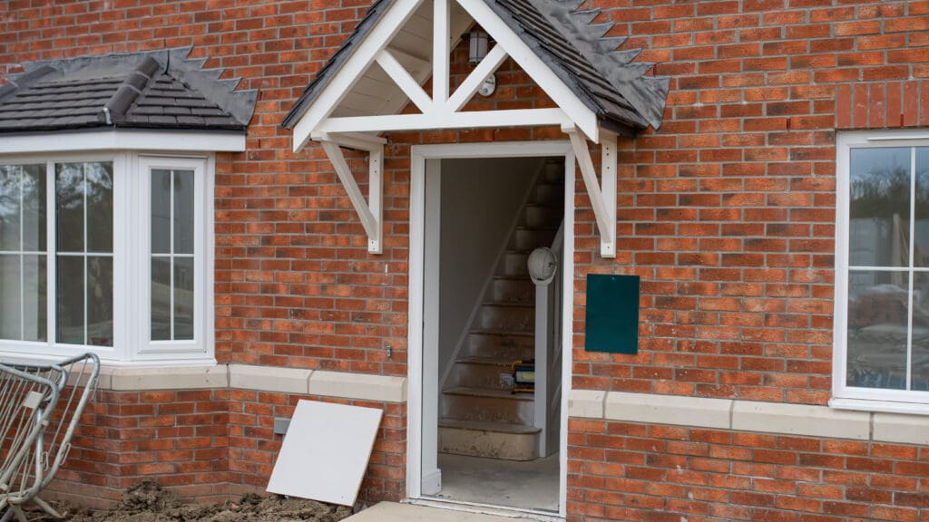 A red brick house with a partially opened front door showing an interior staircase. The entrance has a white wooden awning, and there is a wheelbarrow and construction materials outside, indicating ongoing construction or renovation.