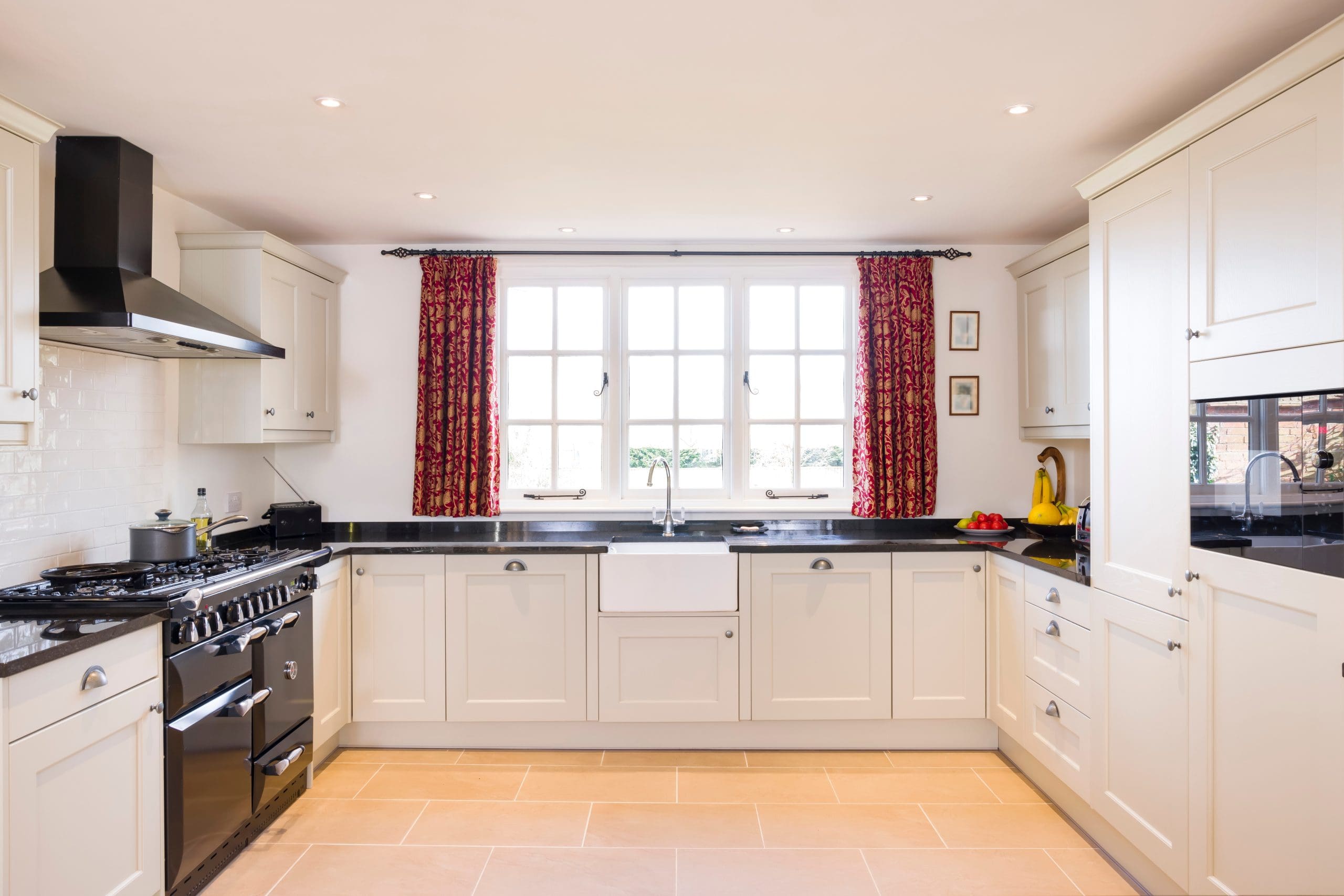 A modern kitchen with white cabinets, black countertops, and stainless steel appliances. A large window with red curtains brings in natural light. There is a white farmhouse sink under the window and light beige tile flooring.