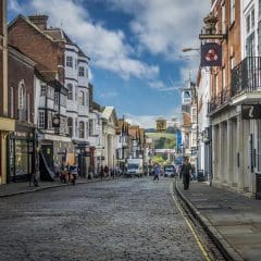 A cobblestone street in a historic town is lined with old buildings featuring storefronts and ornate facades. People can be seen walking and shopping, and a hill with greenery is visible in the background under a partly cloudy sky.