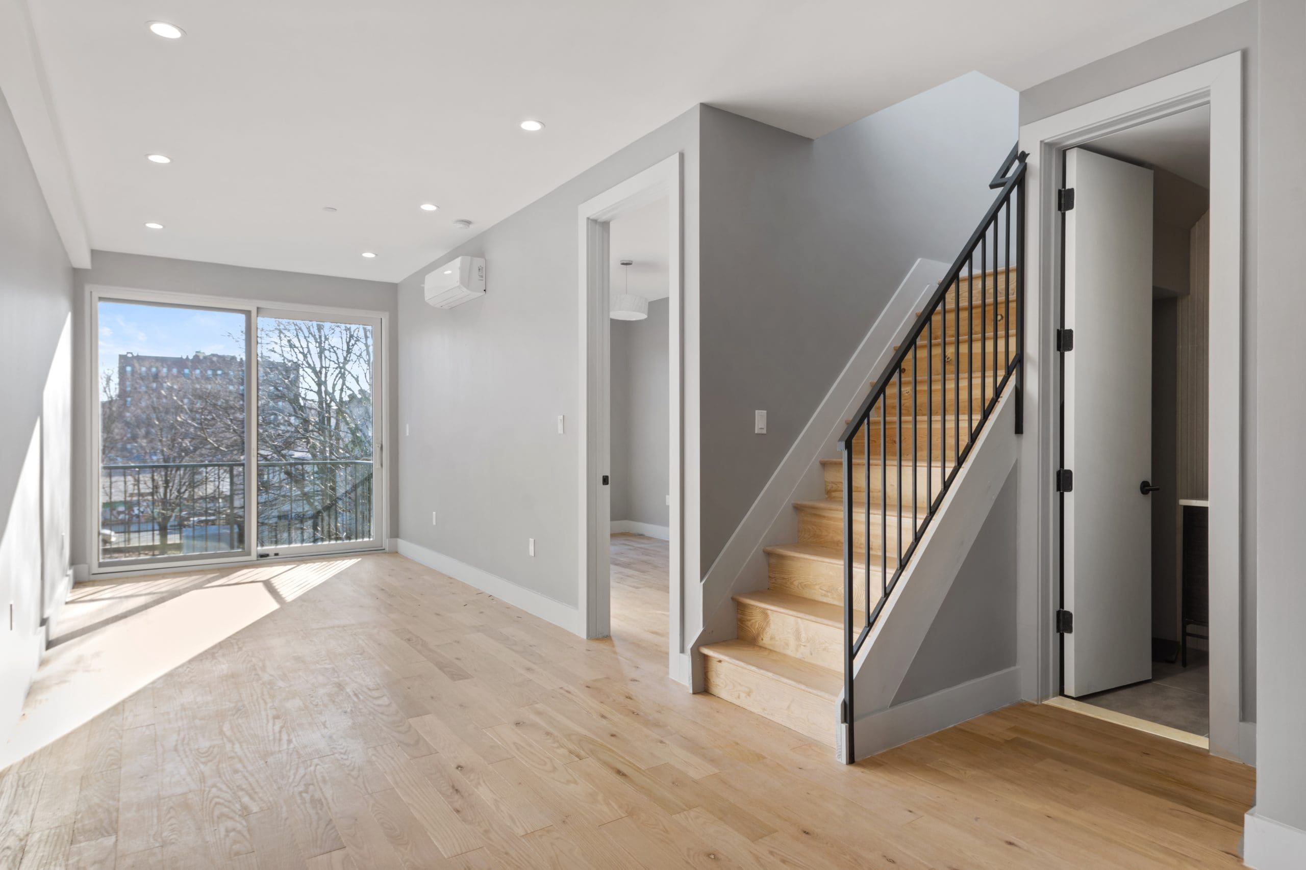 A modern, empty room featuring light hardwood flooring and neutral gray walls. On the right, a wooden staircase with black railings leads up. Large sliding glass doors on the left provide abundant natural light and open to a balcony with a view of trees and buildings.