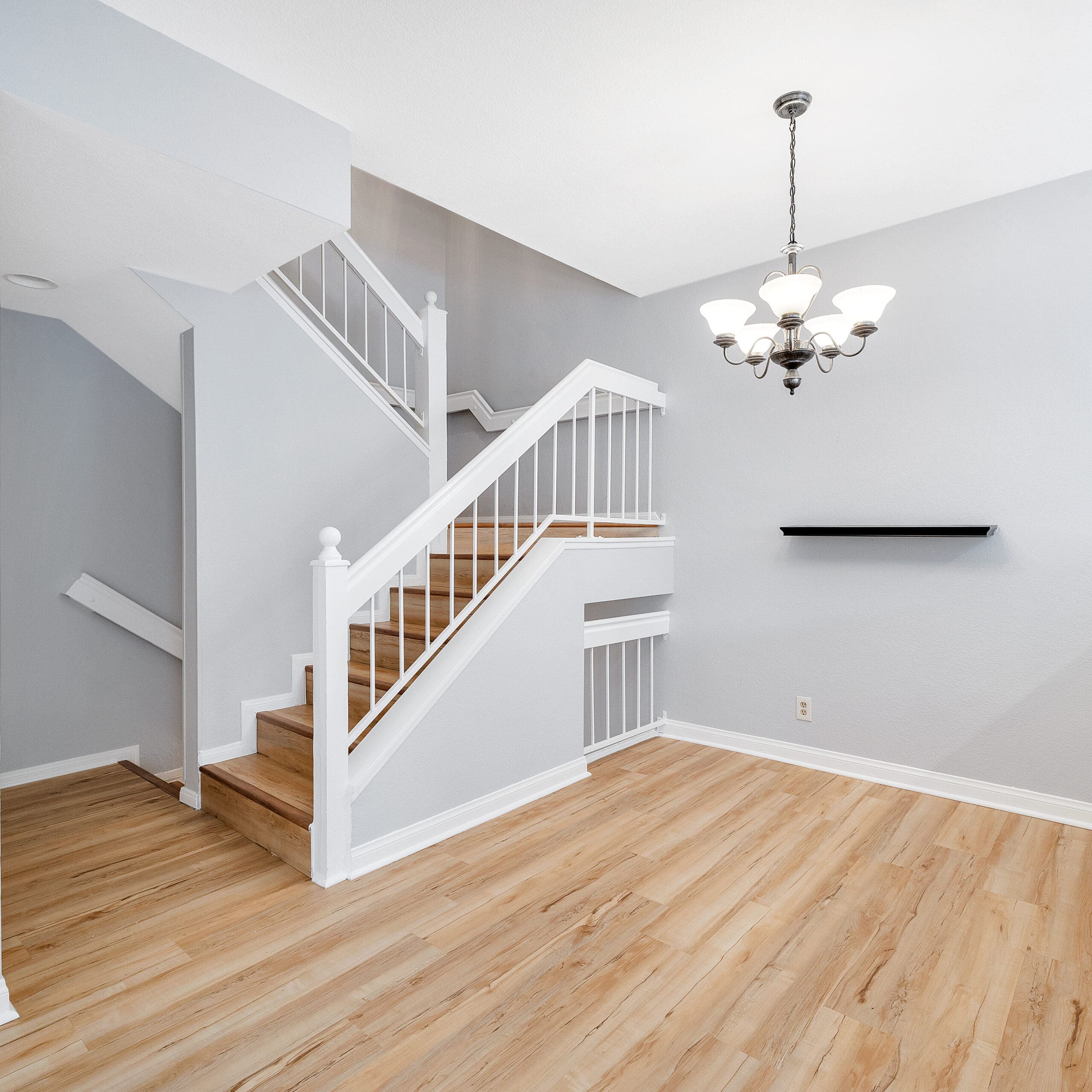 A modern, open-concept interior featuring wooden flooring and light gray walls. A staircase with white railings ascends to the second floor. A black wall shelf and a chandelier with an elegant design adorn the space, adding a touch of sophistication.