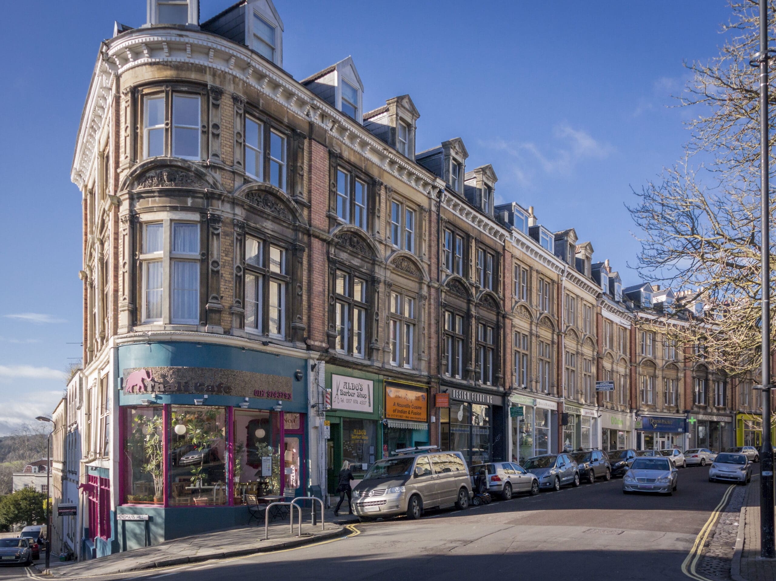 A curved street lined with historic buildings featuring shops and small businesses on the ground floor. Cars are parked along the street, and a sign for "Craft Café" is visible on one building. The sky is clear and trees without leaves are present.