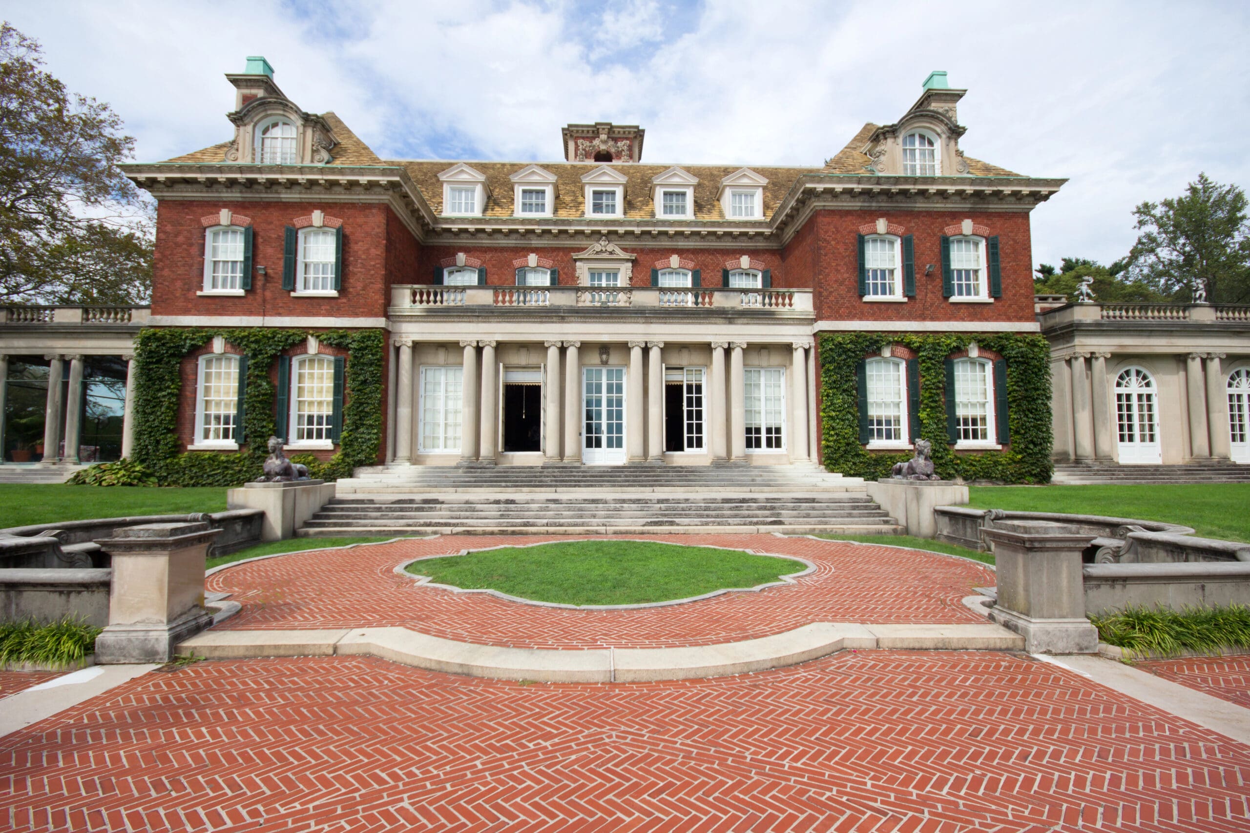 A grand, red-brick mansion with white columns and large windows set in an expansive, landscaped lawn. The front entrance features a semi-circular brick pathway bordered by greenery and statues. Ivy climbs the mansion walls, enhancing its historic elegance.