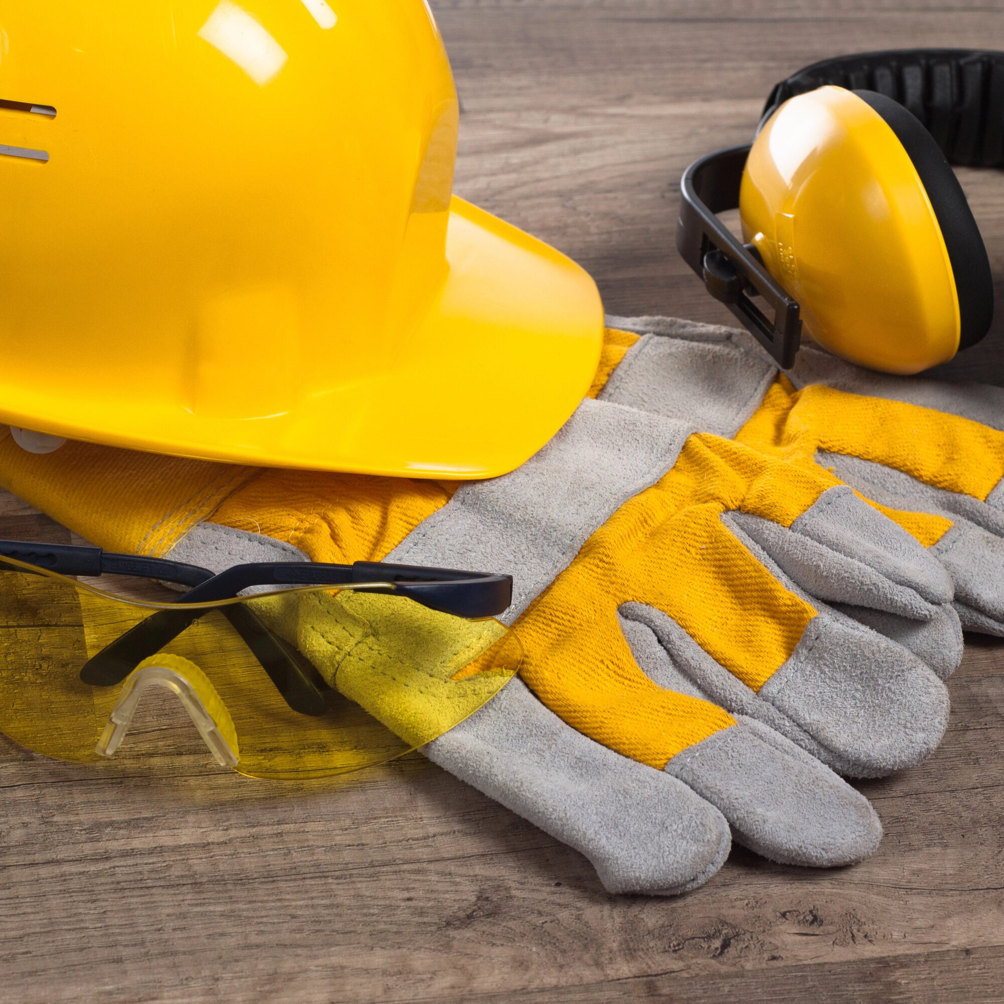 Yellow safety helmet, protective gloves, yellow safety glasses, and ear muffs on a wooden surface. The gloves are gray and yellow, matching the helmet and glasses. The ear muffs are black and yellow.