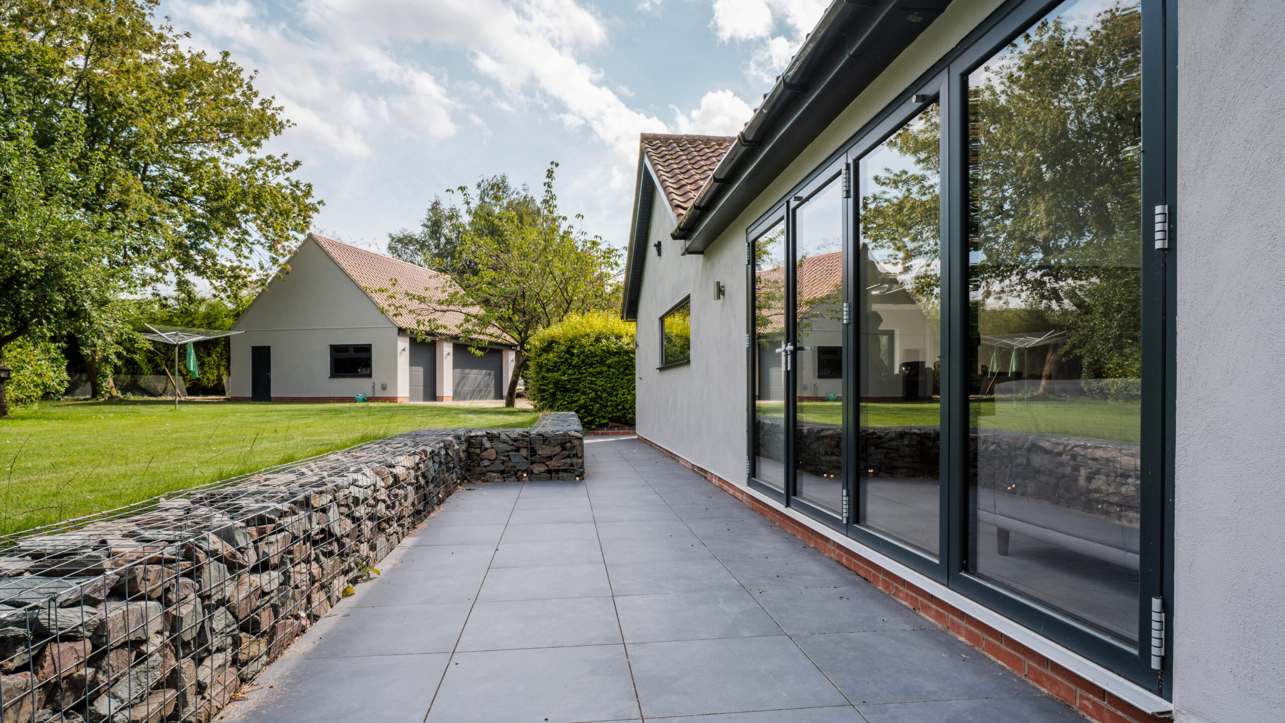 Modern house with large glass sliding doors opening onto a tiled patio, a stone-filled gabion retaining wall, green lawn, trees, and a separate building in the background under a partly cloudy sky.