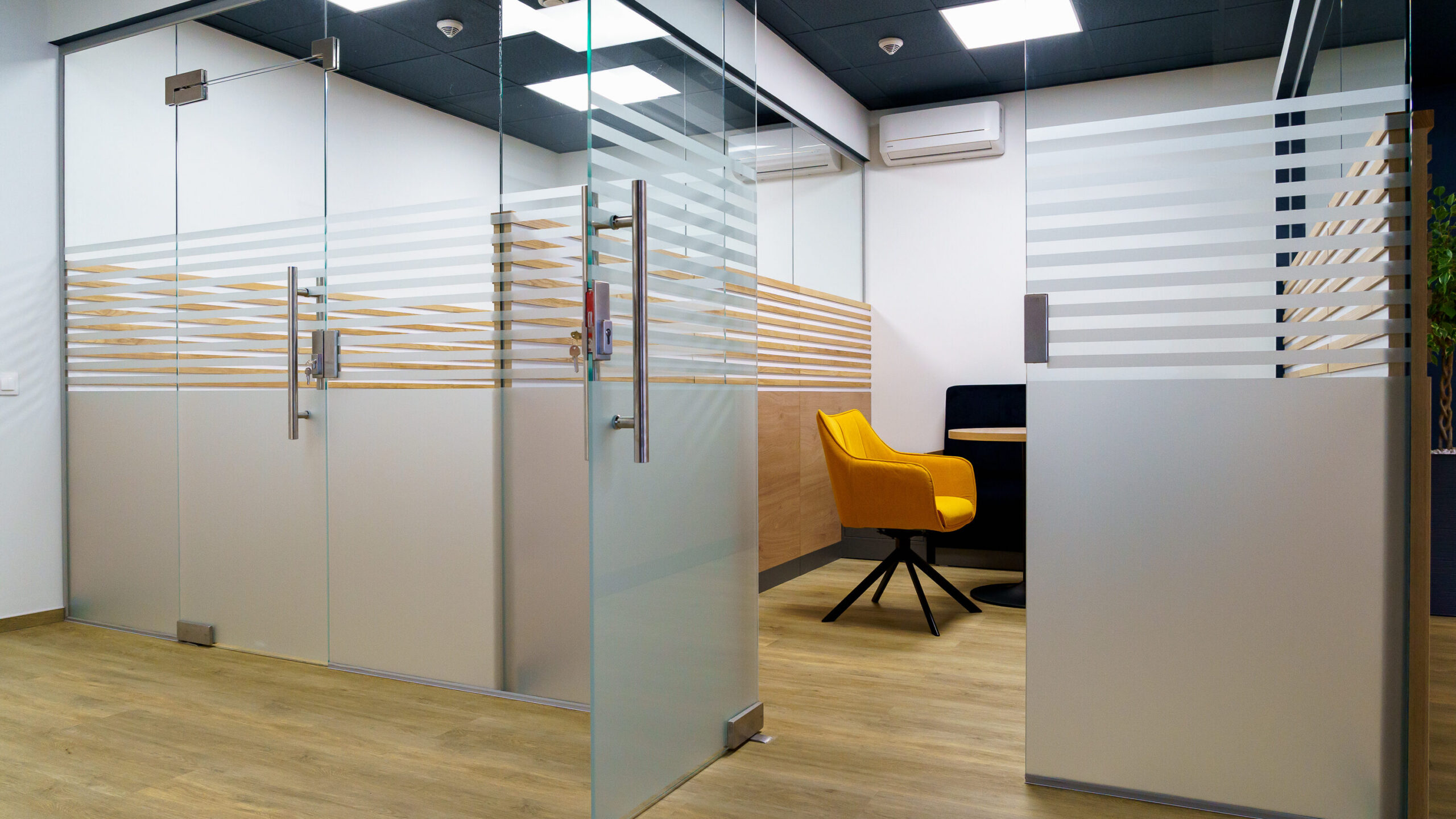 Modern office interior with glass-walled rooms, frosted horizontal stripes on the glass, wood flooring, and a single yellow chair at a desk inside one of the rooms. The space looks clean and minimalist.