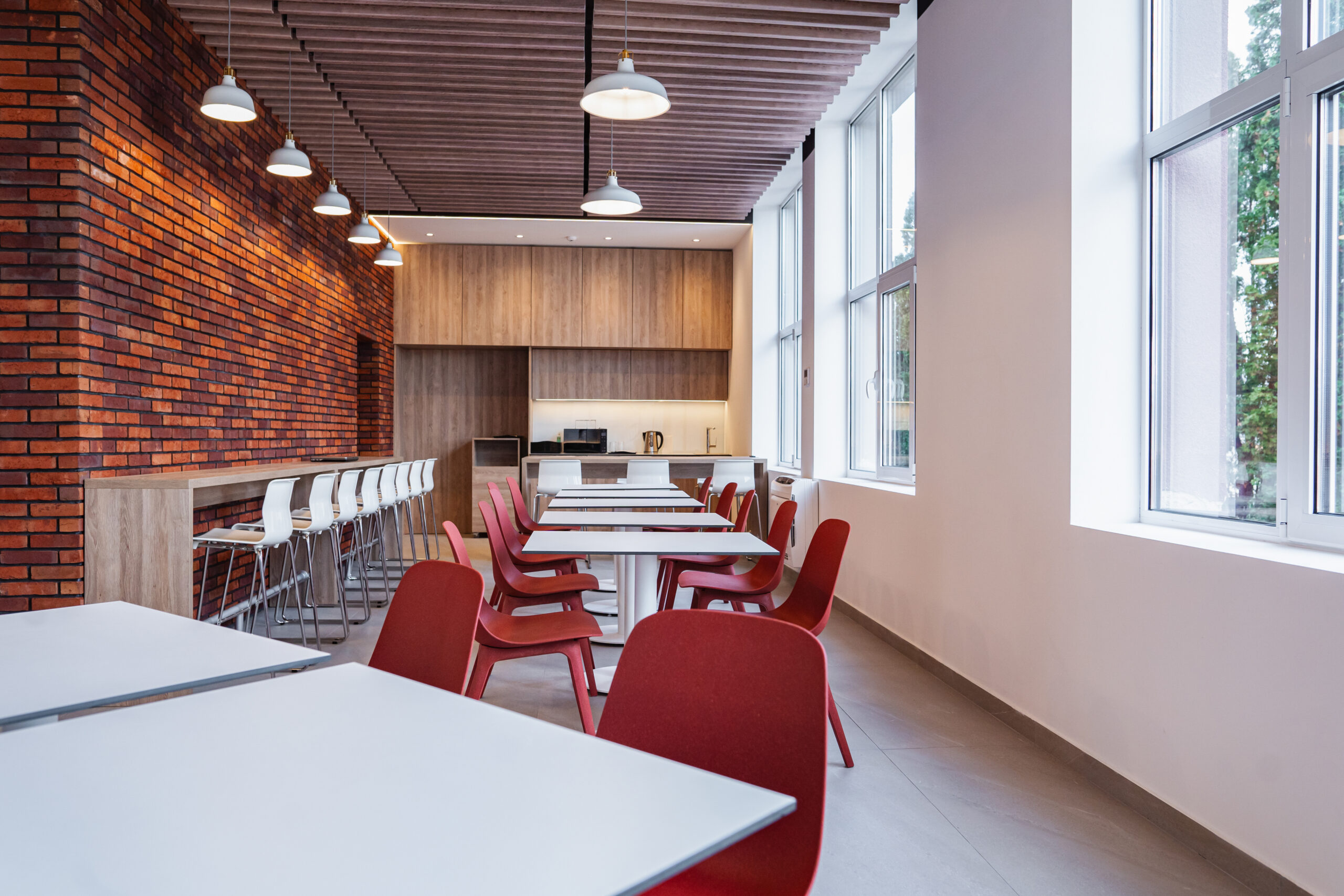 A modern cafeteria with red chairs and white tables, brick accent wall, pendant lights, and large windows letting in natural light. There is a bar counter with high stools and a kitchenette area in the background.