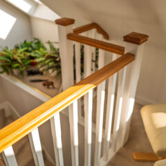A sunlit indoor staircase with white spindles and wooden handrails, leading to a landing with several green potted plants. An upholstered beige chair is partially visible to the right.