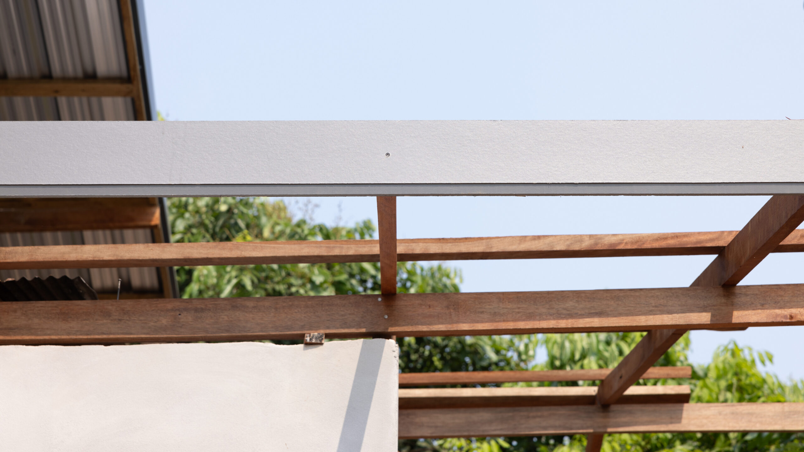 A close-up view of a wooden roof frame under construction with a metal beam, set against a background of green trees and a clear blue sky.