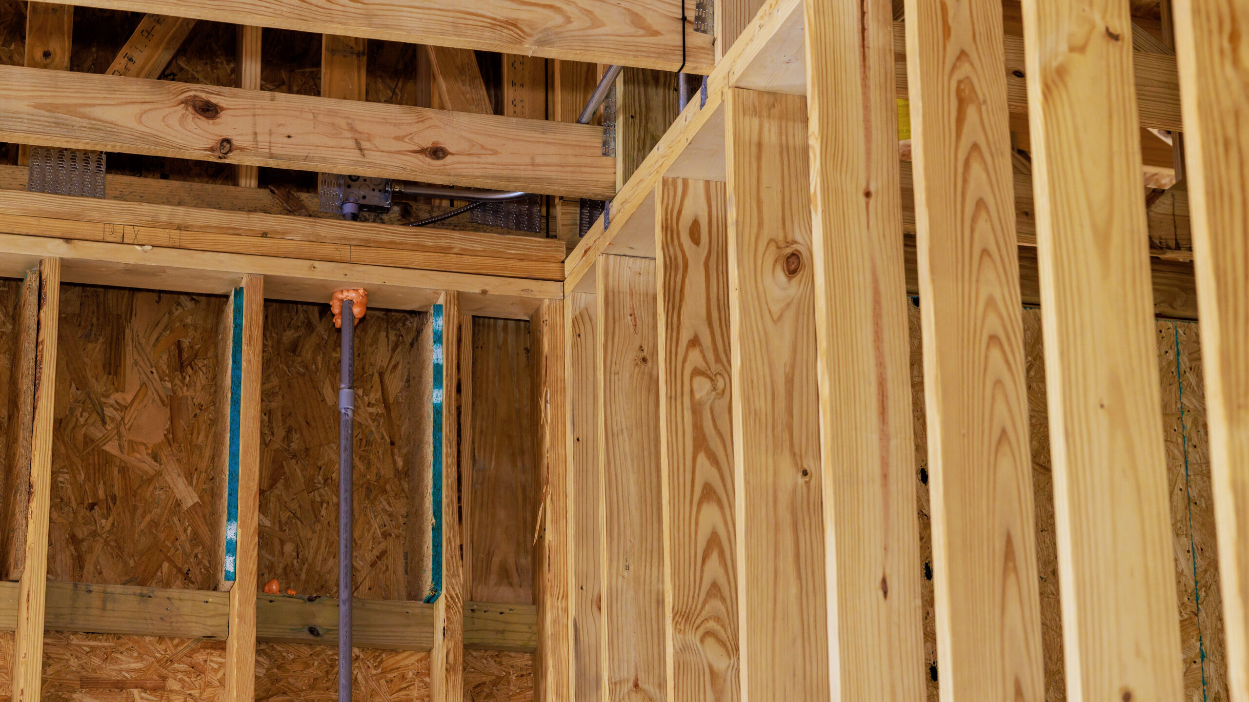 Wooden framing and beams of a house under construction, showing exposed studs, joists, and unfinished plywood walls. A pipe runs vertically through the frame, indicating rough plumbing installation.