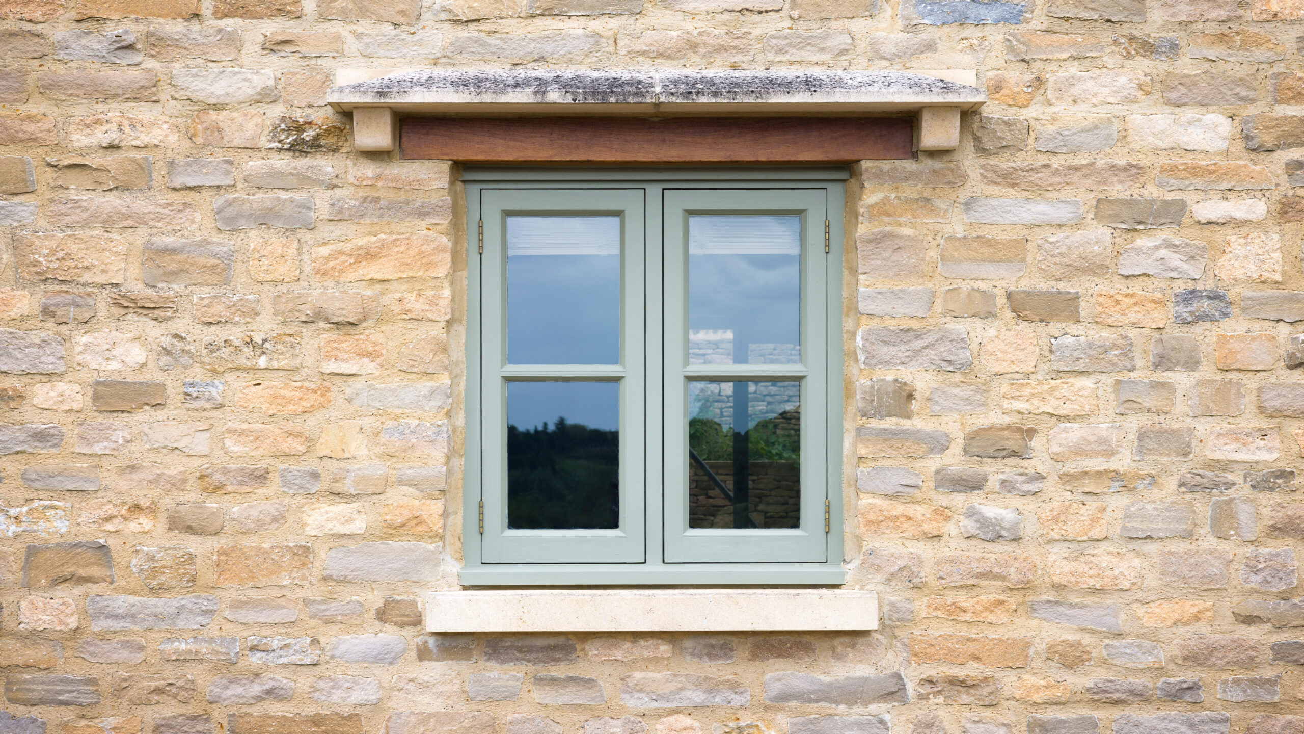 A green-framed window with four glass panes set in a stone wall, reflecting trees and sky from outside. The window has a stone sill and a wooden lintel above it.
