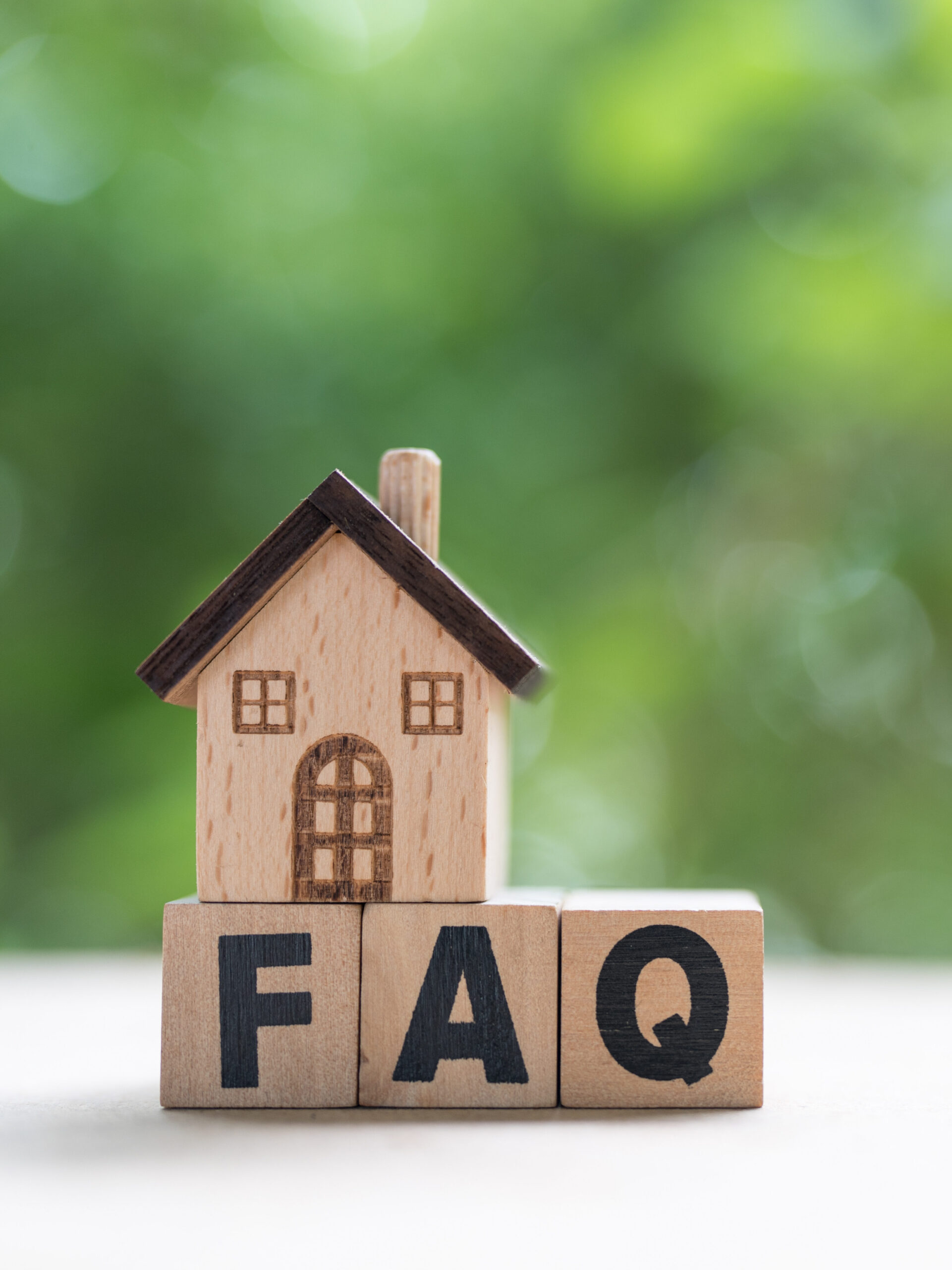 A small wooden house model sits atop three wooden blocks displaying the letters "FAQ" against a blurred green background.