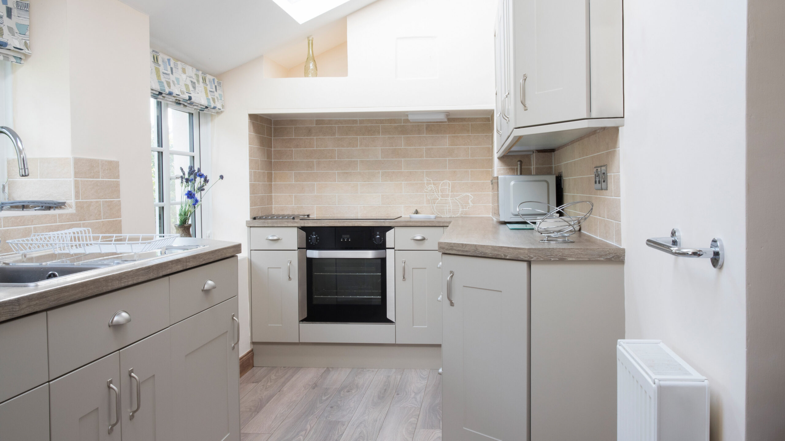Bright, modern kitchen with light cabinetry, built-in oven, beige subway tile backsplash, gray countertops, a window with patterned curtains, and wooden flooring. A sink, microwave, and dish rack are visible.