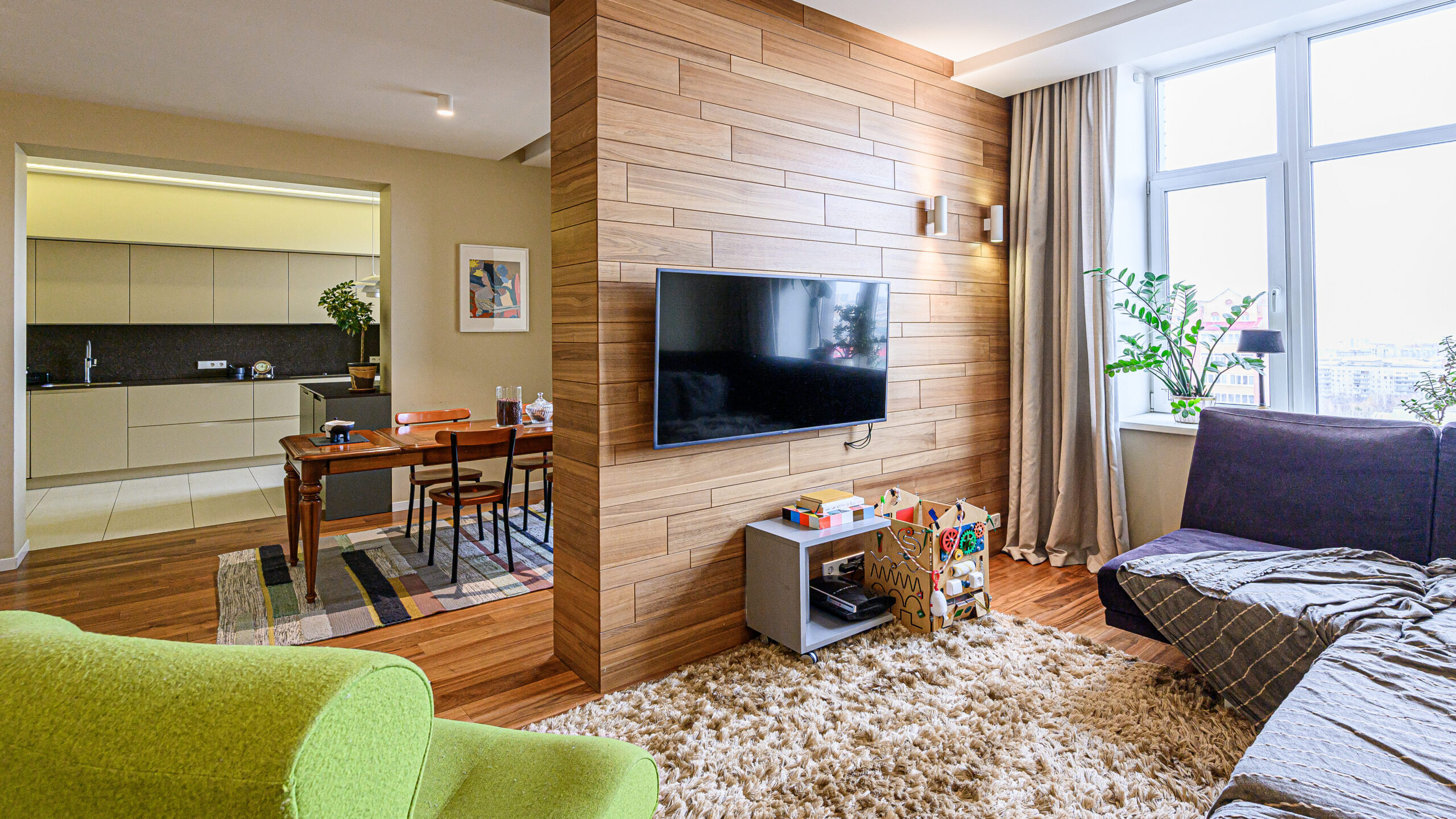 A modern apartment living room with a wood-paneled wall, mounted TV, cozy seating, a play area with toys, a rug, and an open dining area with a table and chairs. Large window lets in natural light.