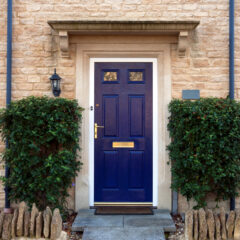 A navy blue front door with a brass mail slot and handle is set in a light stone house, flanked by two large green bushes and a black wall lantern. A low stone border lines the small entrance path.