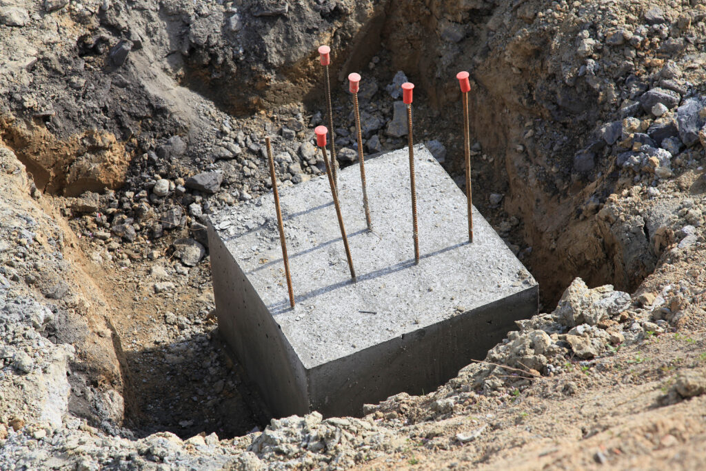 A concrete foundation block with several protruding steel rebar rods capped with red safety covers is set in a dirt excavation site surrounded by soil and rocks.