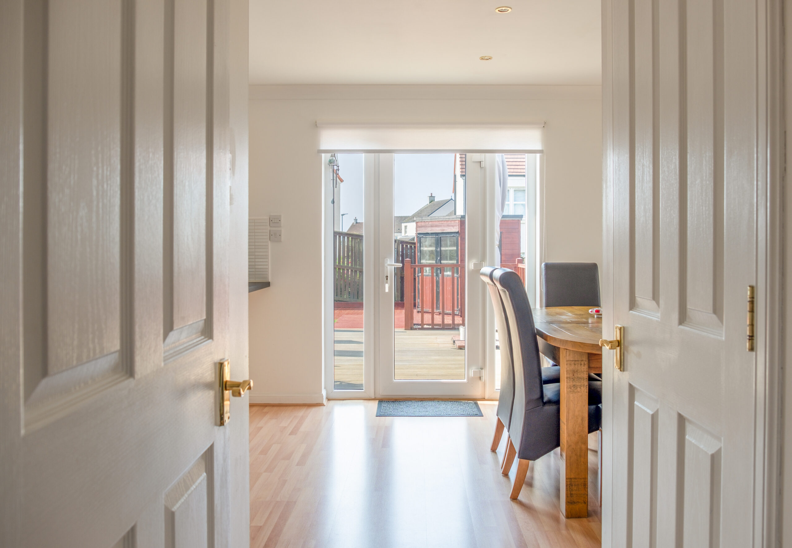 Bright dining area with wooden floors and a table with chairs, seen through open double doors. Large glass doors at the back let in natural light and offer a view of a fenced yard and neighboring houses.