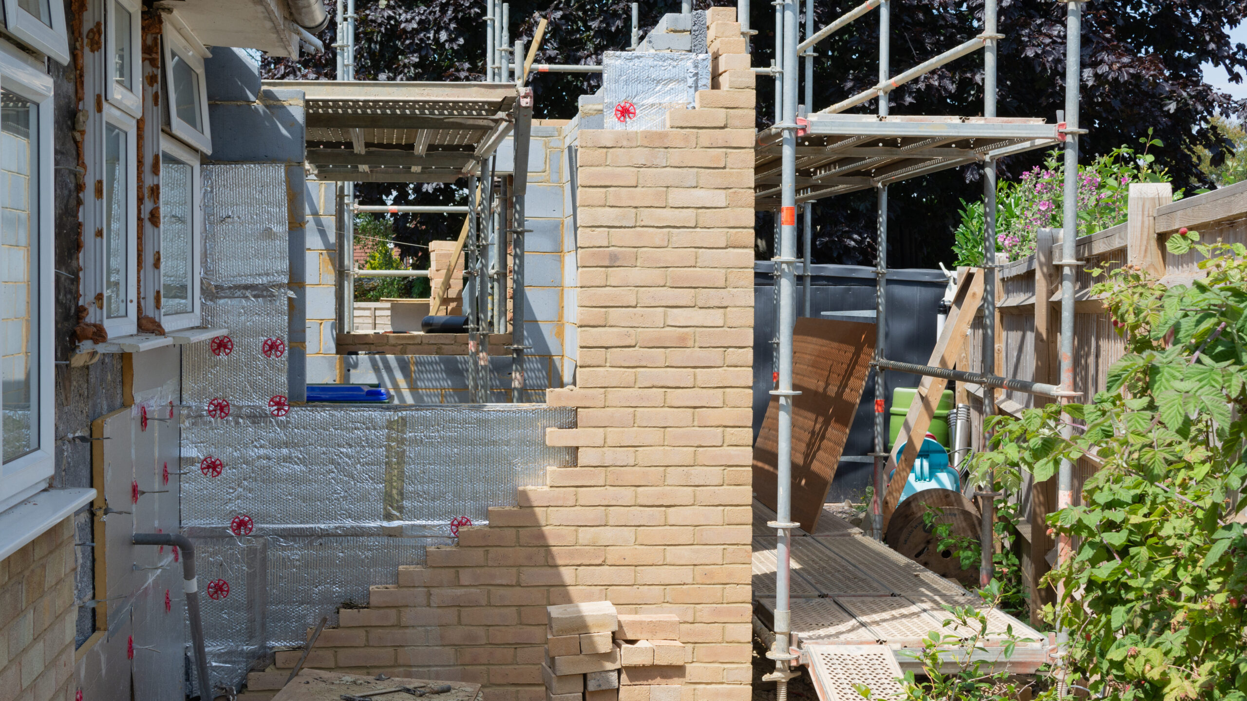 A partially built brick wall with scaffolding around it, insulation panels on the left, and garden plants on the right, showing ongoing construction work on a house extension.