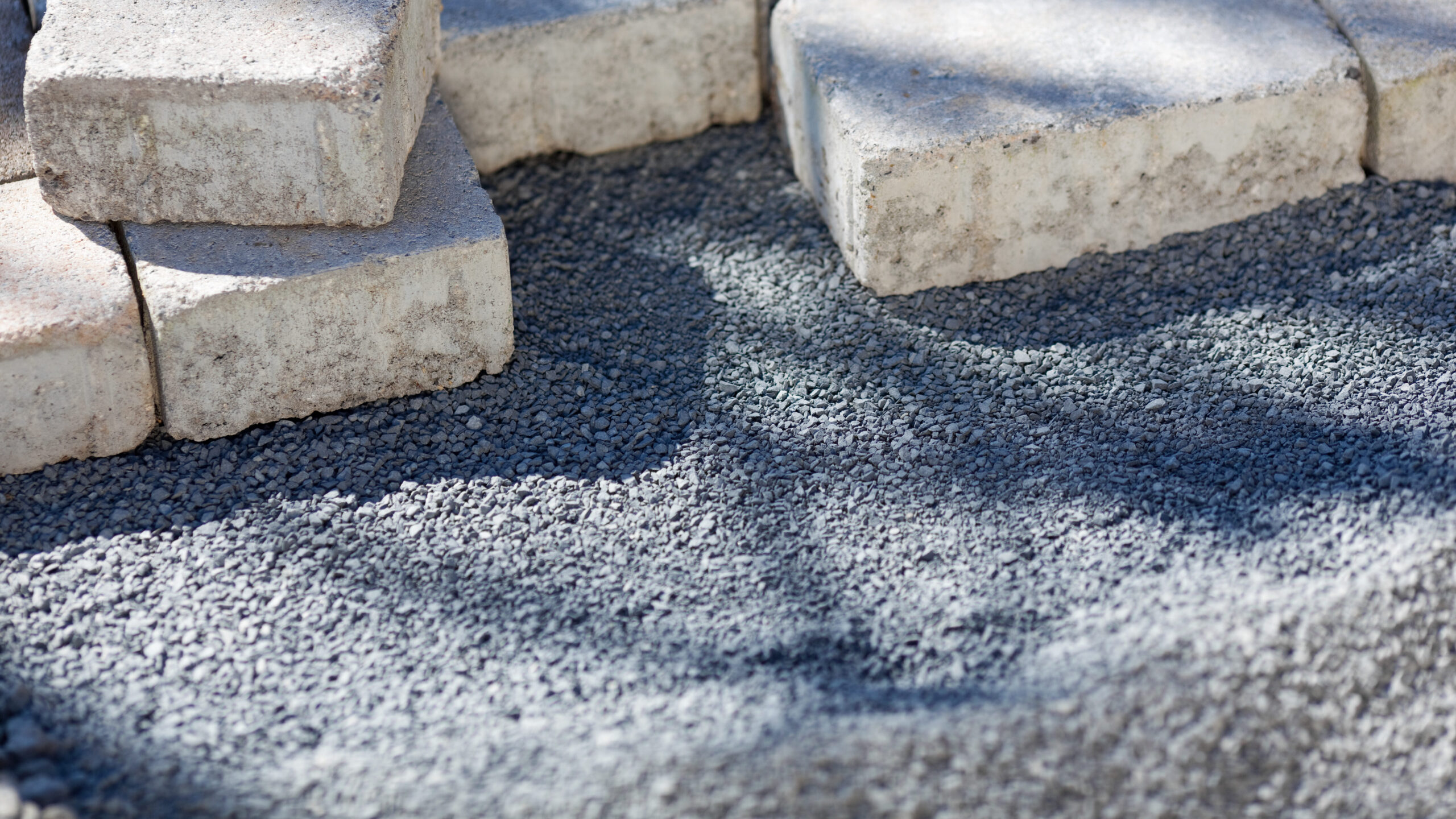 Close-up of several light-colored paving stones placed on a bed of gray gravel, ready to be installed for a landscaping or construction project. Shadows from nearby objects fall across the gravel.
