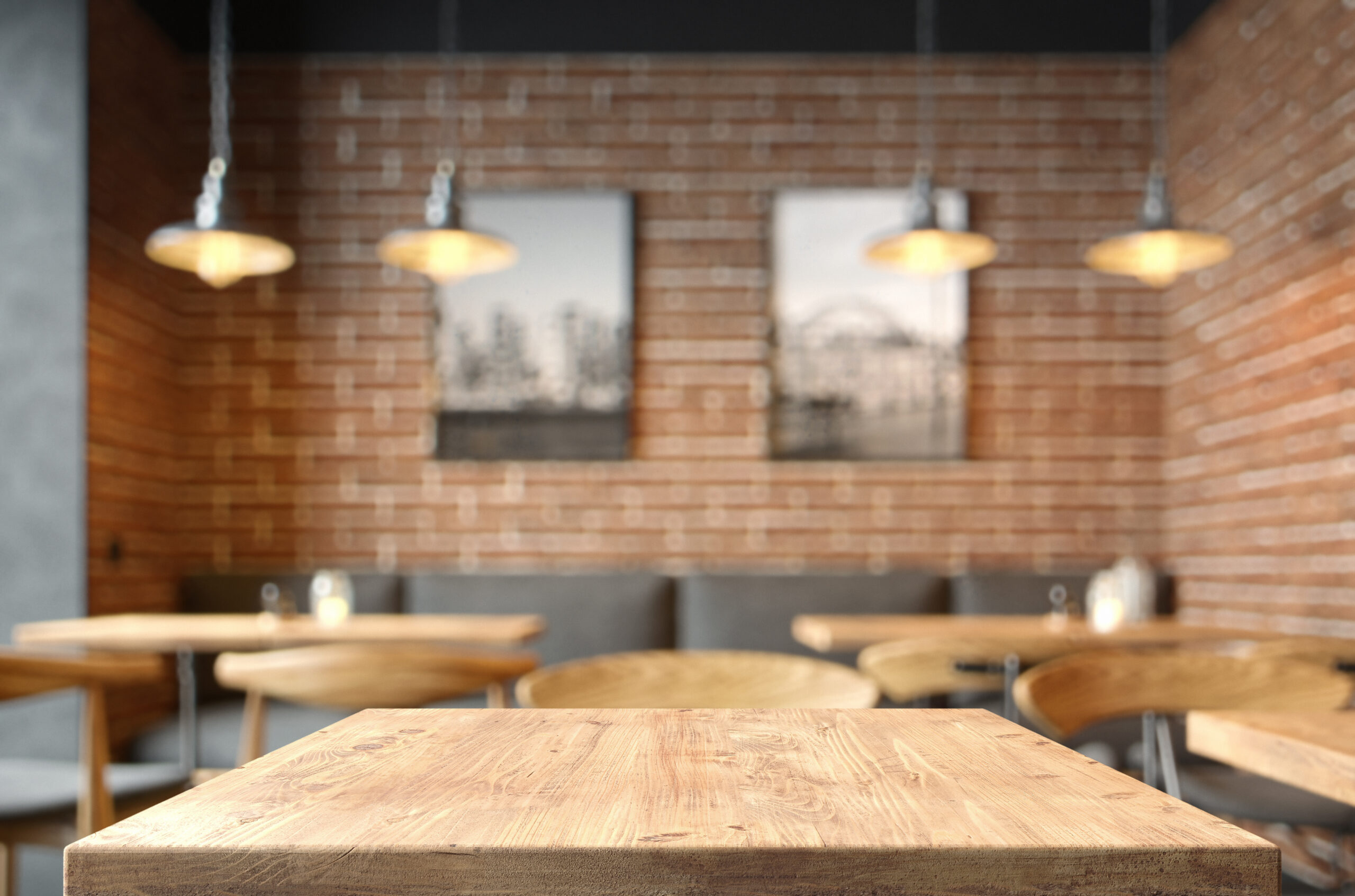 A close-up of a wooden table in a cozy cafe with brick walls, hanging pendant lights, and blurred tables, chairs, and black-and-white photos in the background.