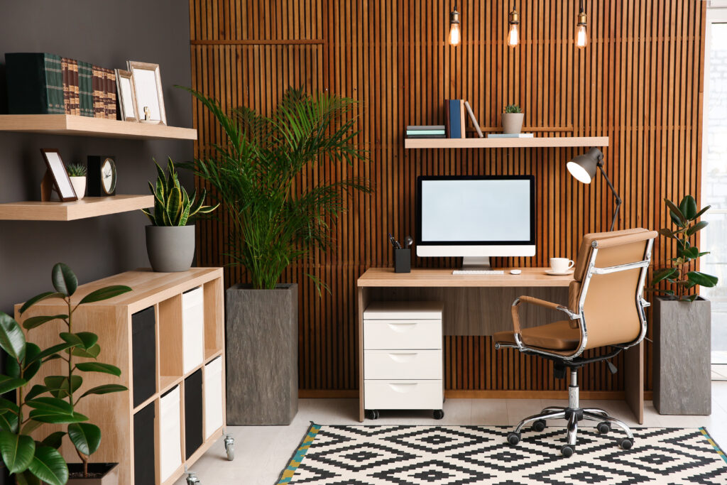A modern home office with a wooden desk, computer, swivel chair, large green plants, floating shelves with books and decor, a geometric rug, and vertical wooden wall panels with hanging lights.