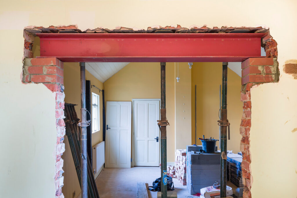 A partially demolished wall with exposed bricks and a red steel beam supported by metal props, inside a room under renovation with yellow walls and white double doors in the background.