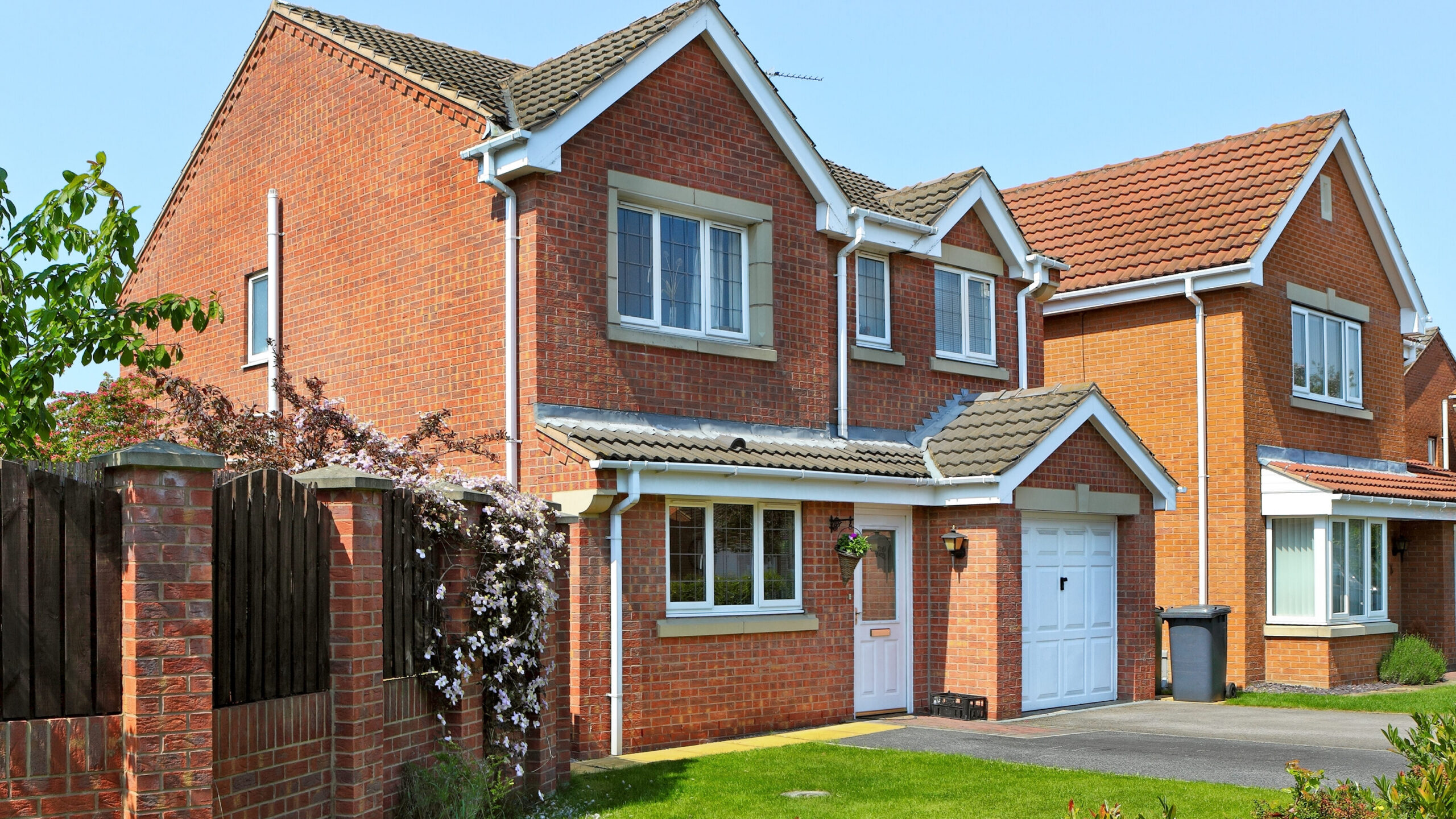 A modern two-story red brick house with a white garage door, white-framed windows, and a small front garden, set on a sunny day with clear blue skies.