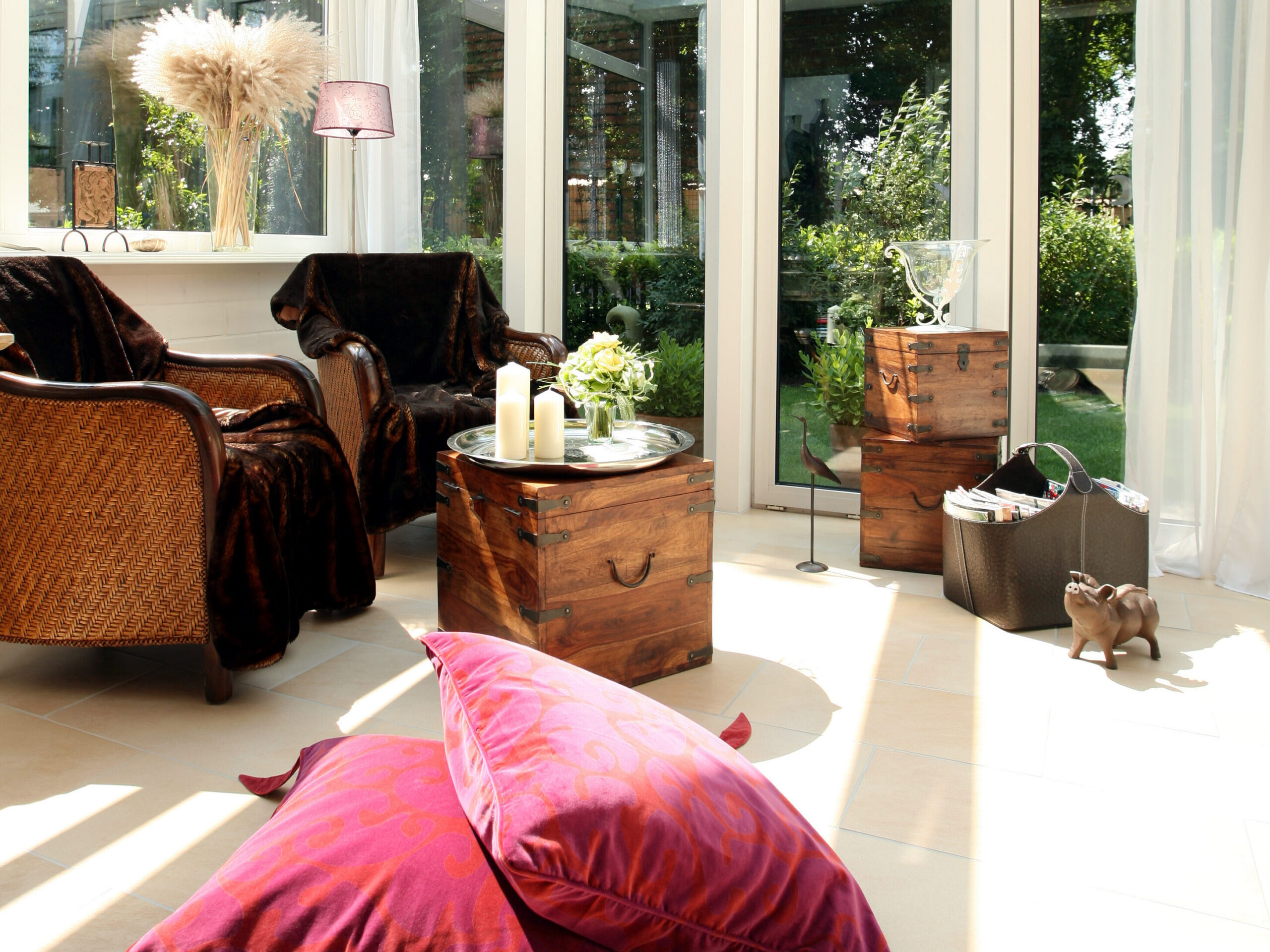 A sunlit room with wicker chairs draped in dark blankets, wooden chests as tables, candles, a glass vase, red cushions on the floor, and large windows showing a green garden outside.