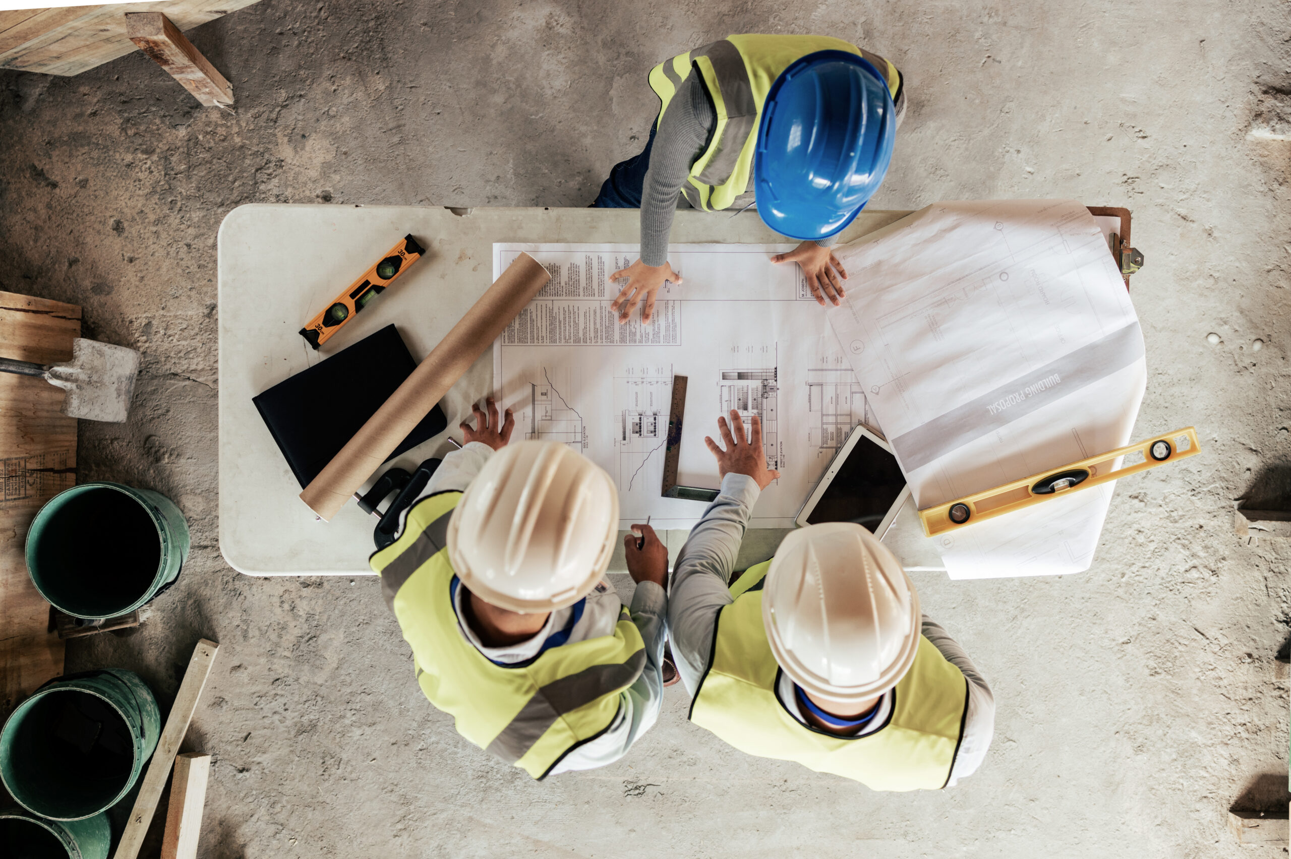 Three construction workers in safety vests and helmets review blueprints and documents on a table at a construction site, with tools and a tablet nearby.